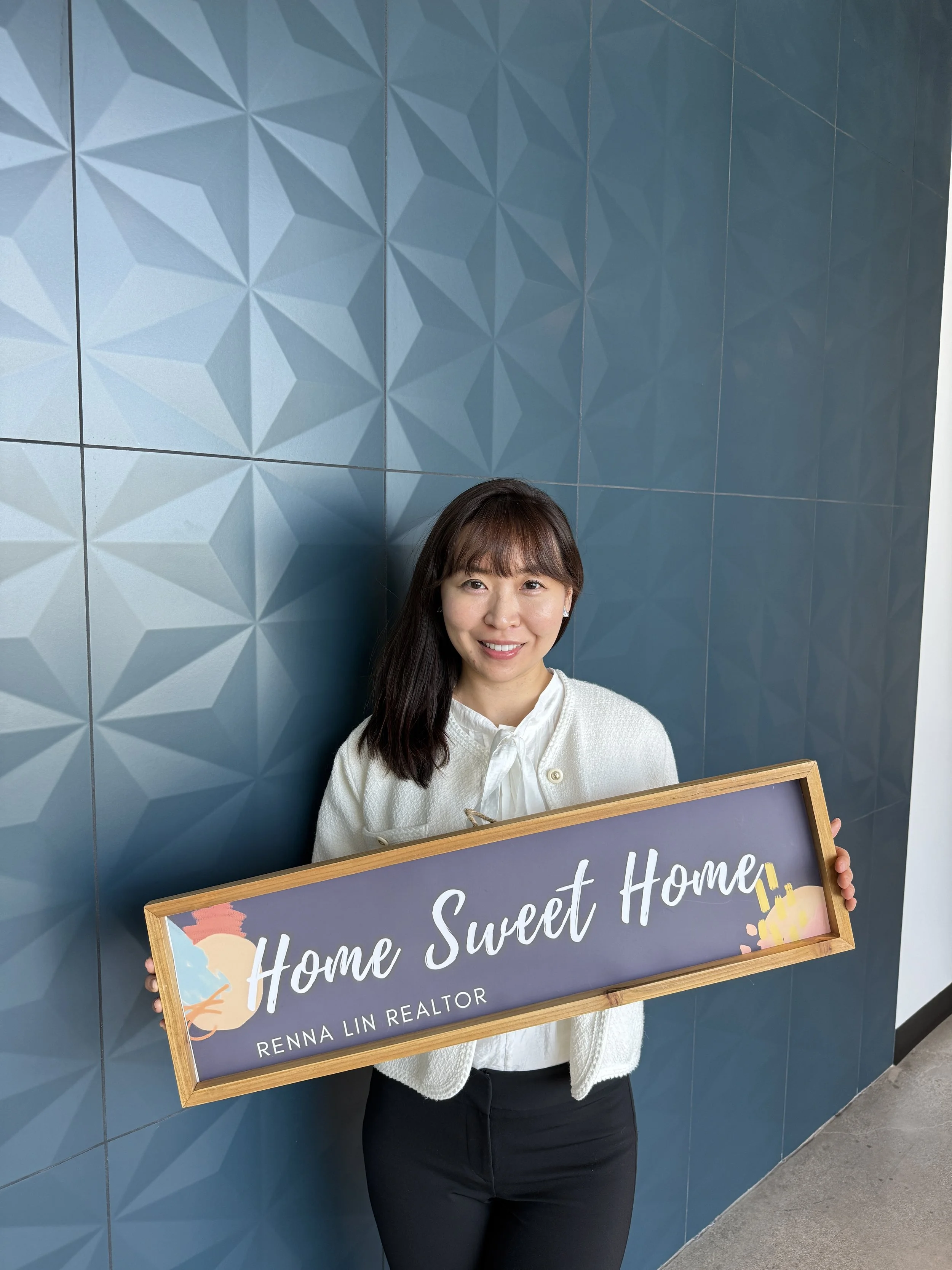 A woman with long dark hair and bangs smiling, standing against a blue geometric wall, holding a framed sign that reads 'Home Sweet Home'.