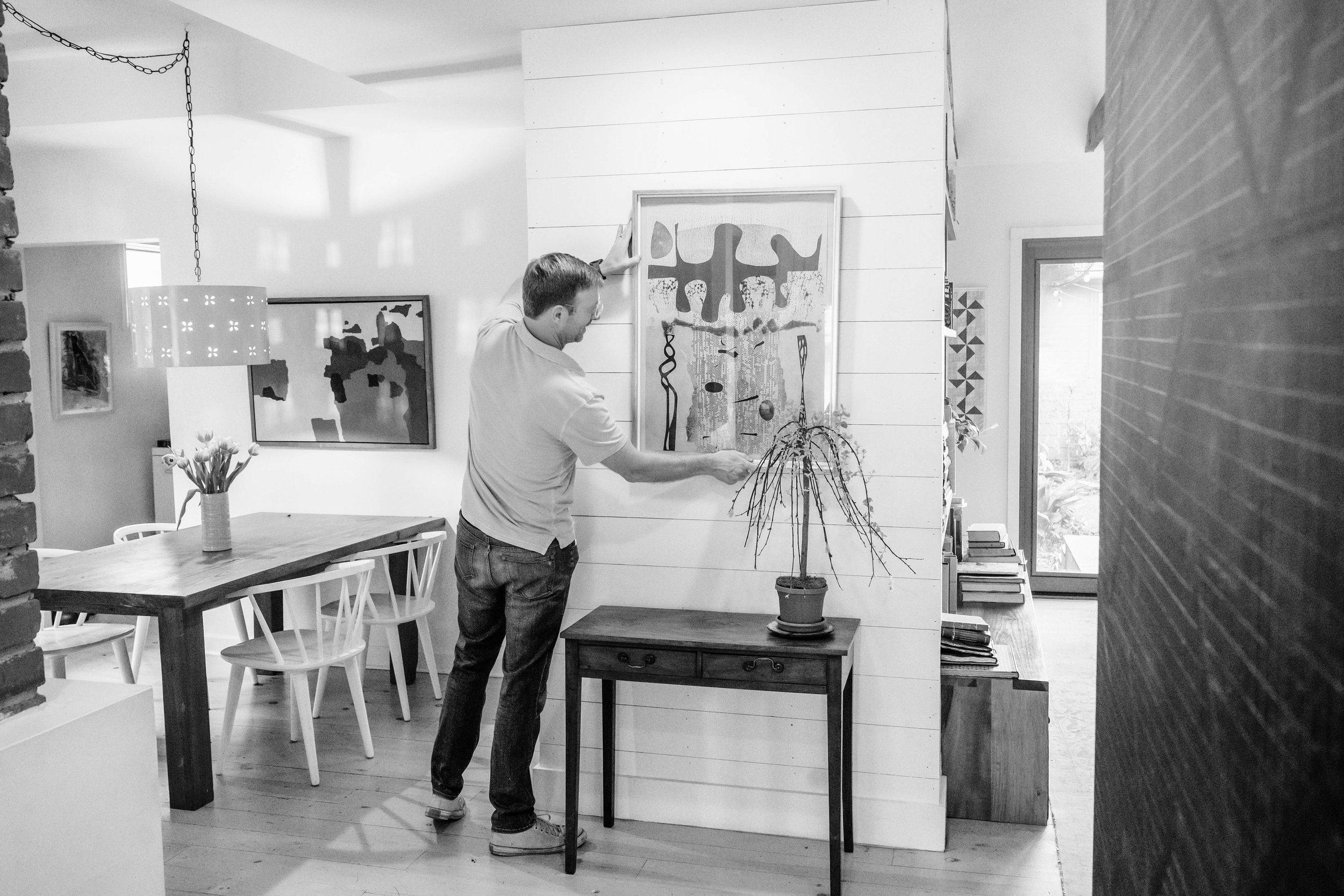 A man hangs a framed abstract painting on a white wooden-paneled wall in a modern home interior.