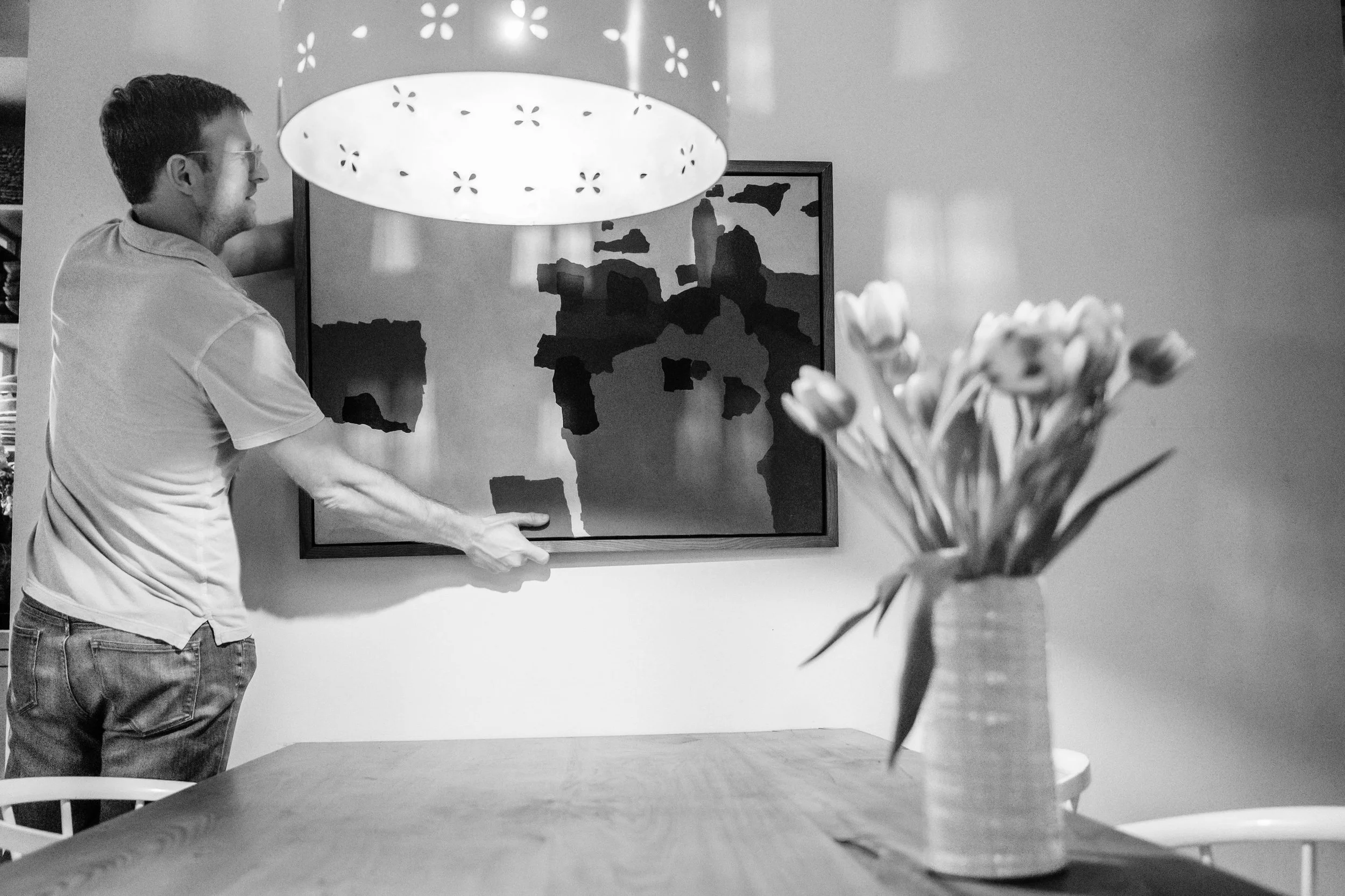 A man hanging a framed abstract art piece on a wall in a dining area, with a vase of tulips on the table in the foreground.