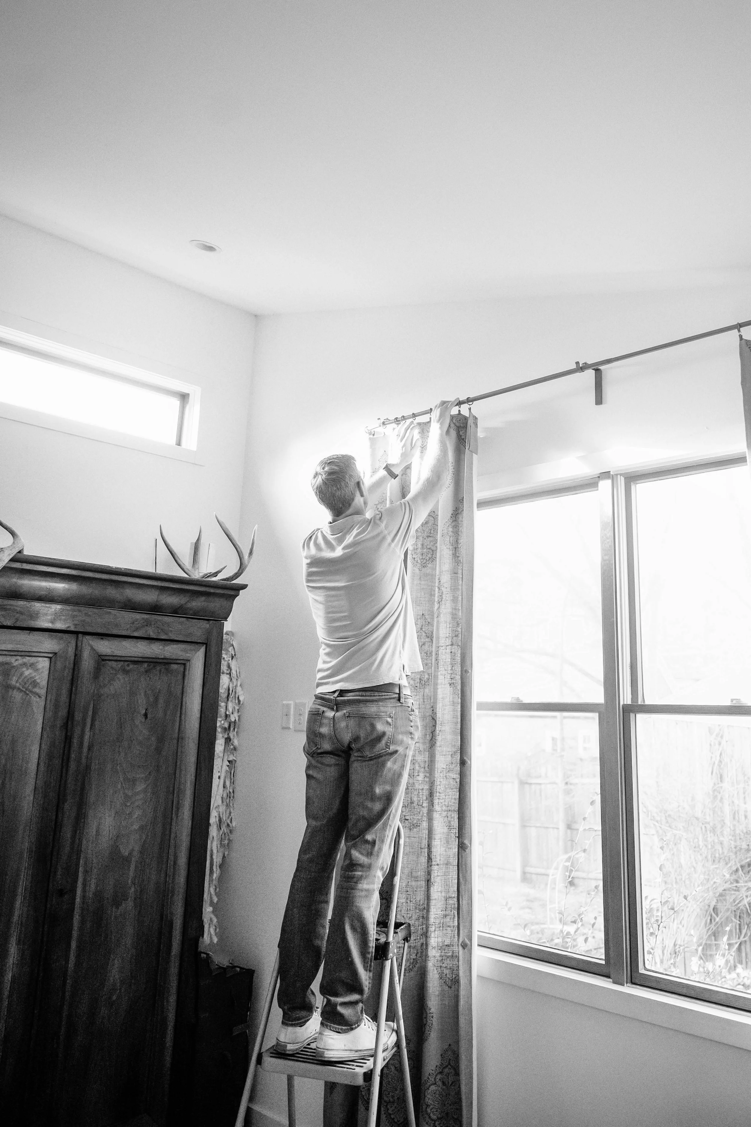 A person standing on a step stool, reaching up to hang curtains on a window rod inside a room, with natural light coming through large windows.