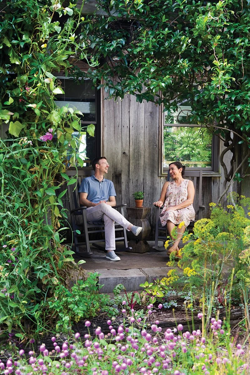 A man and woman sitting on a porch surrounded by lush green plants and flowers, smiling and talking to each other.