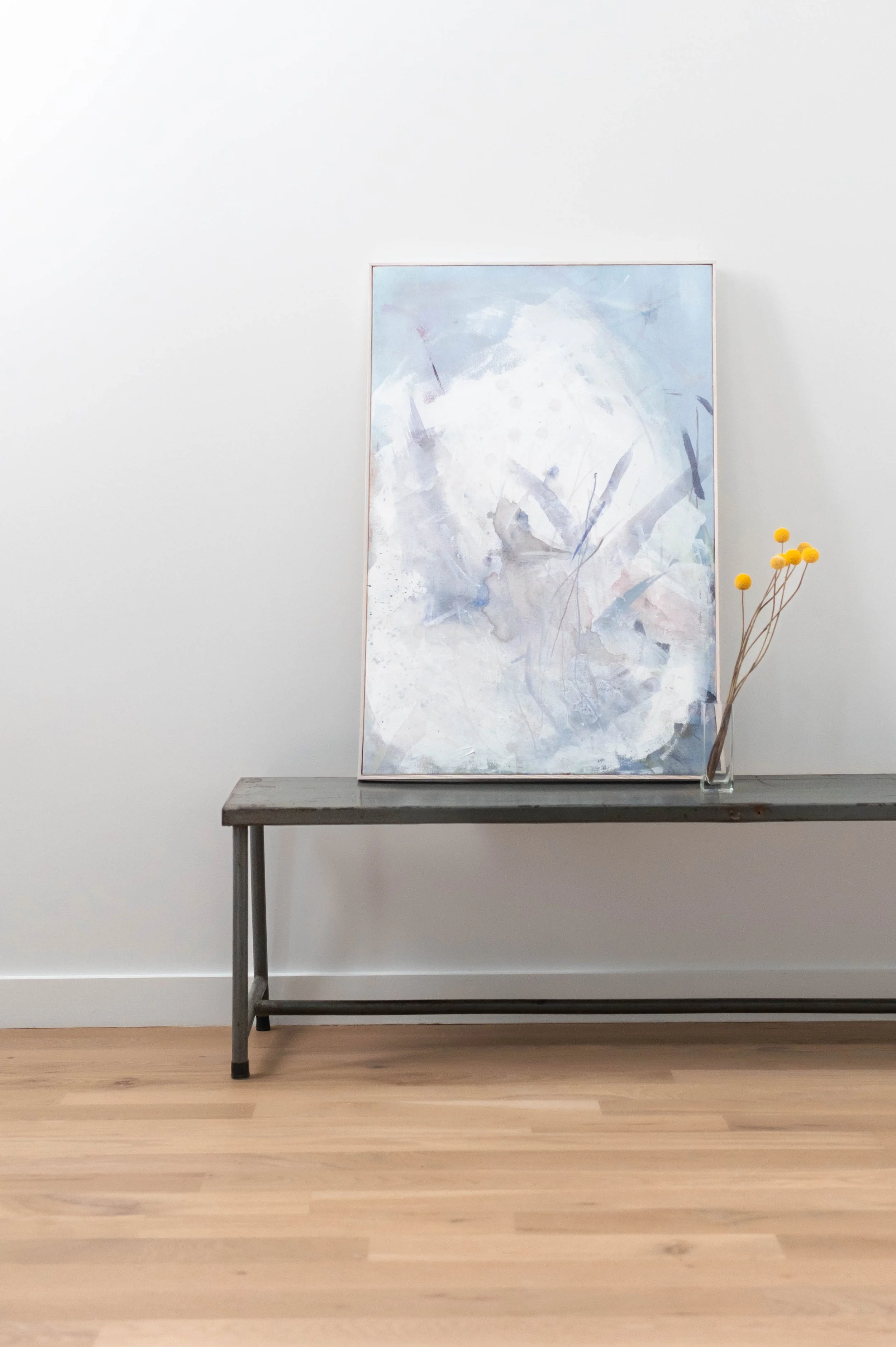 An abstract painting in soft blue and white tones on a wooden bench, with a small glass vase holding yellow pom-pom flowers next to it, against a plain white wall.