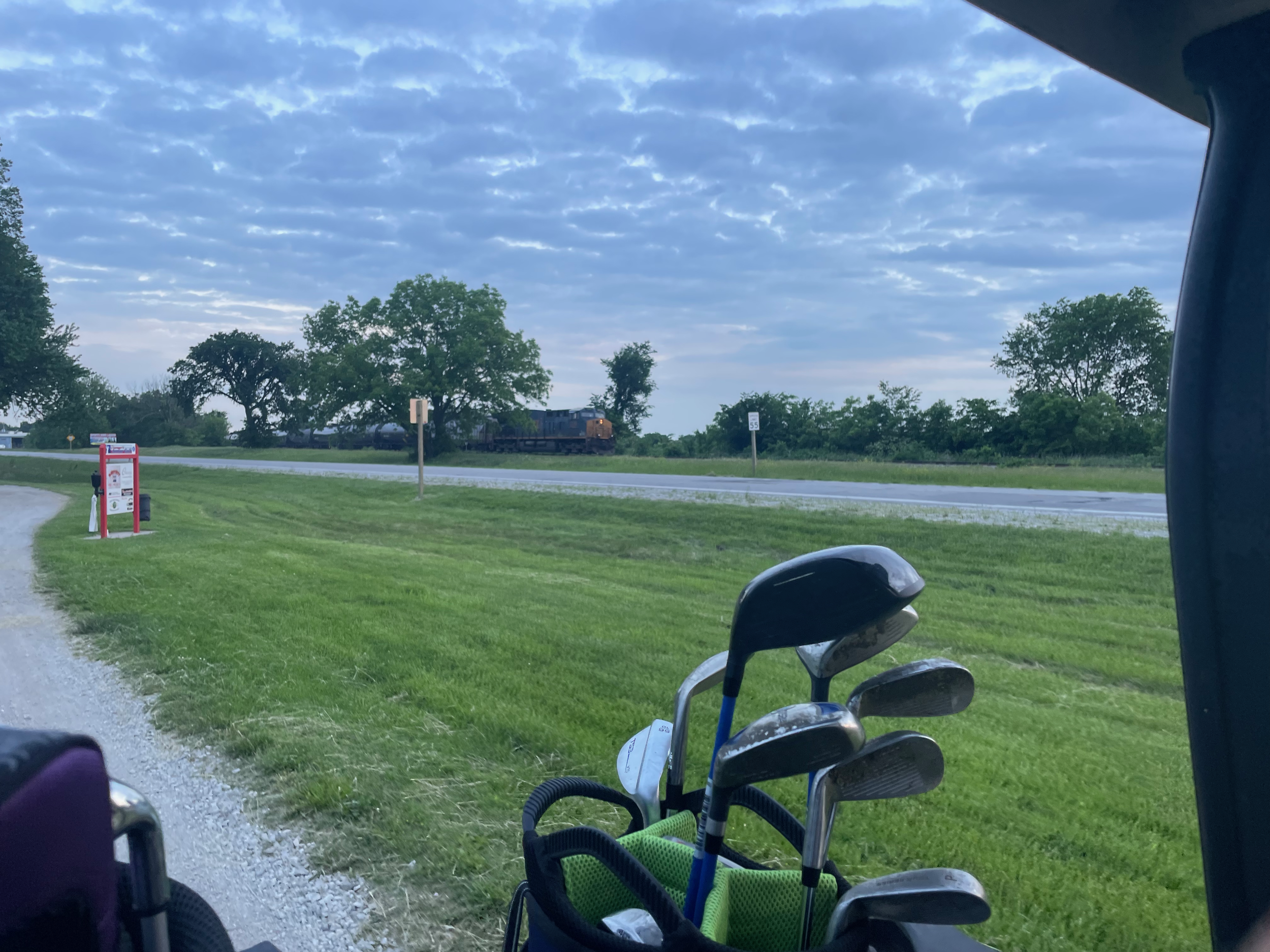 Golf bags with clubs in a golf cart on a grassy area near a small road. In the background, a train is visible moving along the tracks under a partly cloudy sky.