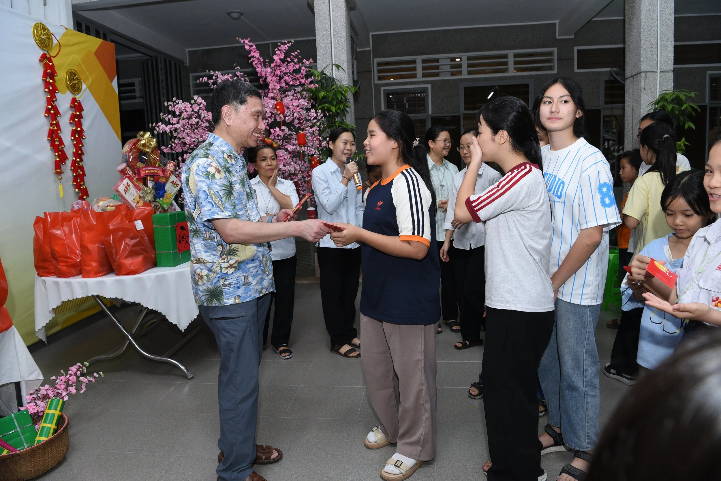 A woman receiving a red envelope from a man in a floral shirt during a celebration, with a line of women and girls waiting behind her. Decorations include a cherry blossom tree, red banners, and gift bags.