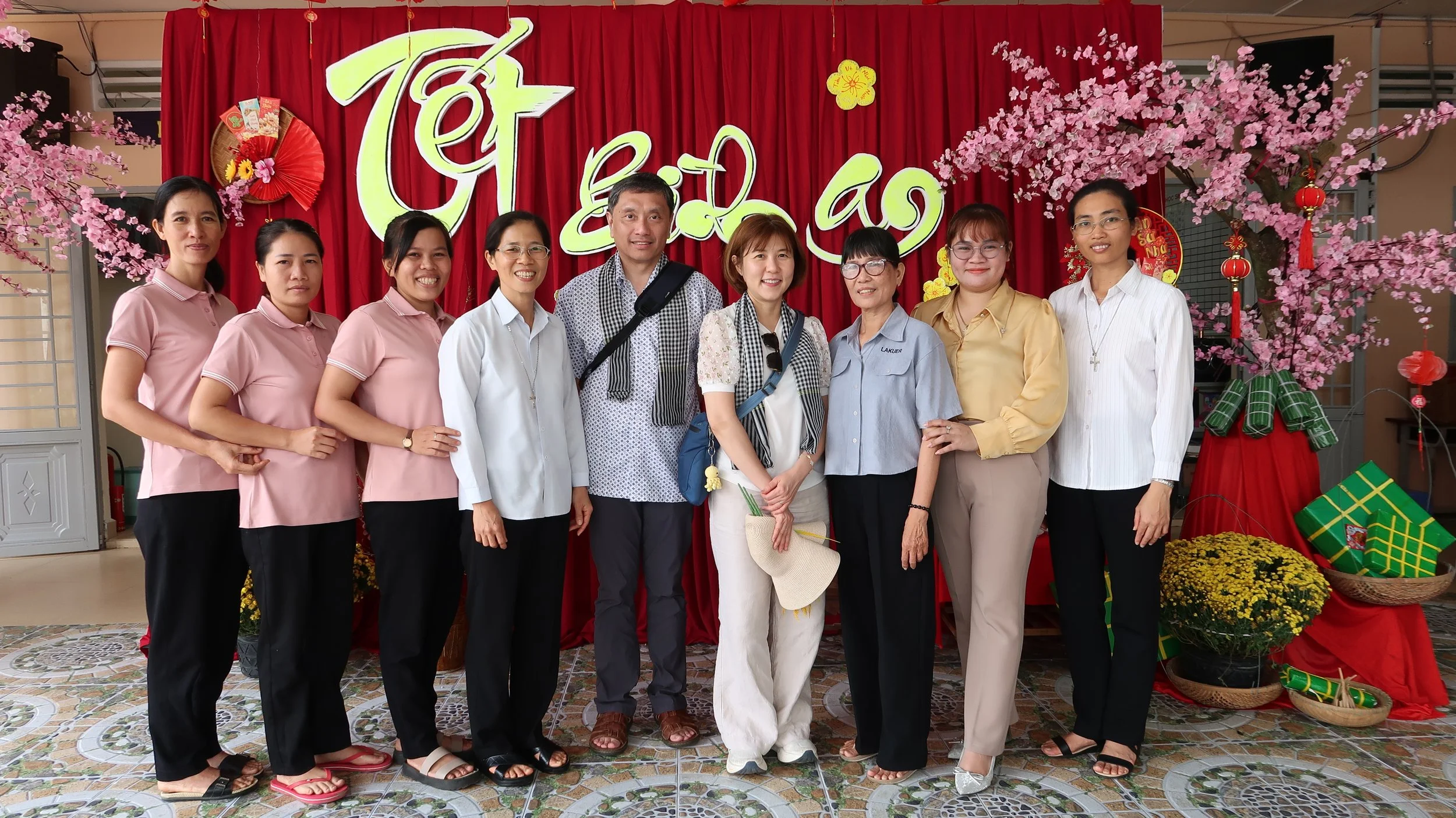 A group of ten people standing in front of red and pink decorations with cherry blossom flowers for a celebration. The background features red hanging lanterns, gift boxes, and yellow flowers.