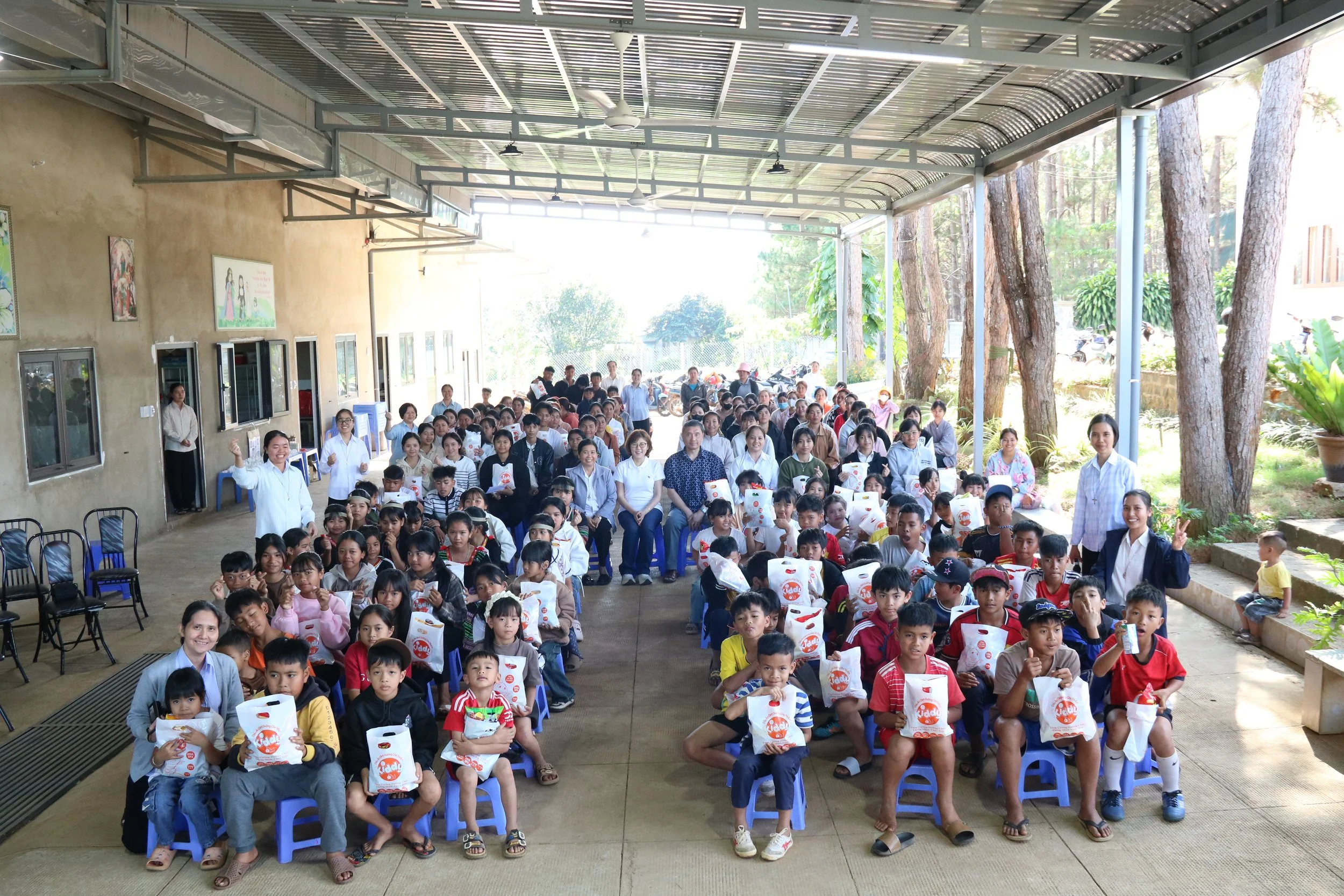 A large group of children and adults gathered in a covered outdoor area, some children holding white bags, with trees and greenery in the background.