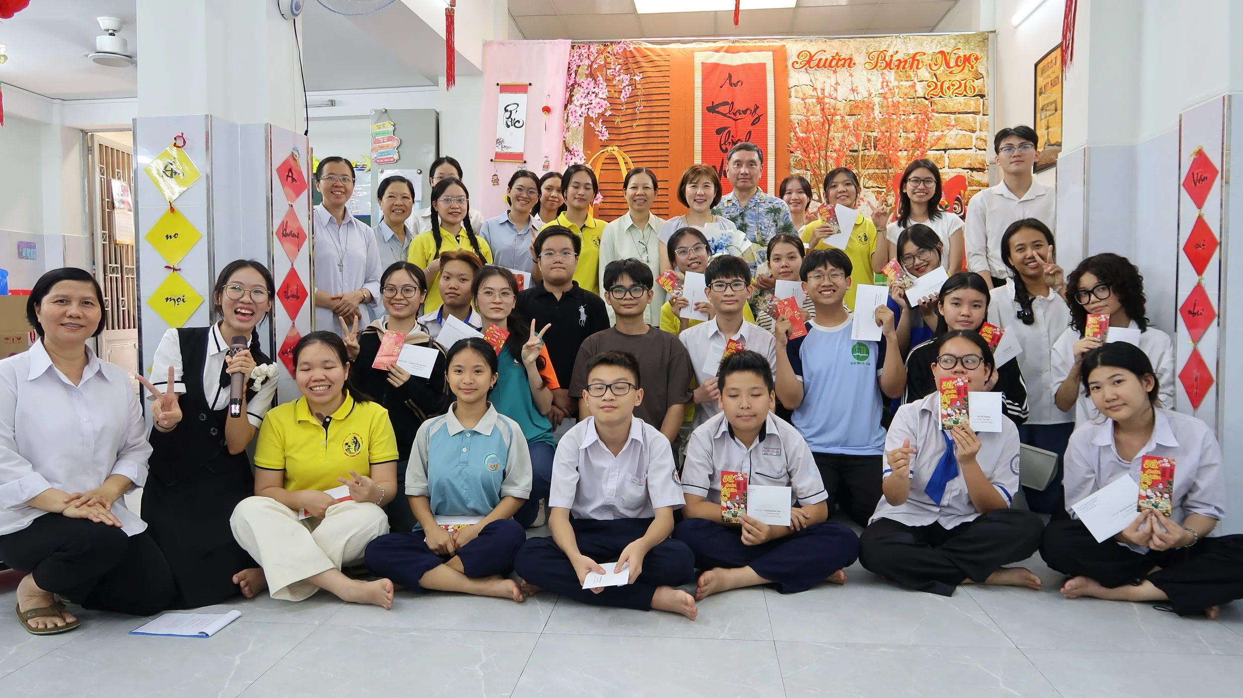 Group of children and adults gathered in a decorated room, celebrating an event, holding cards and envelopes, smiling for the photo.