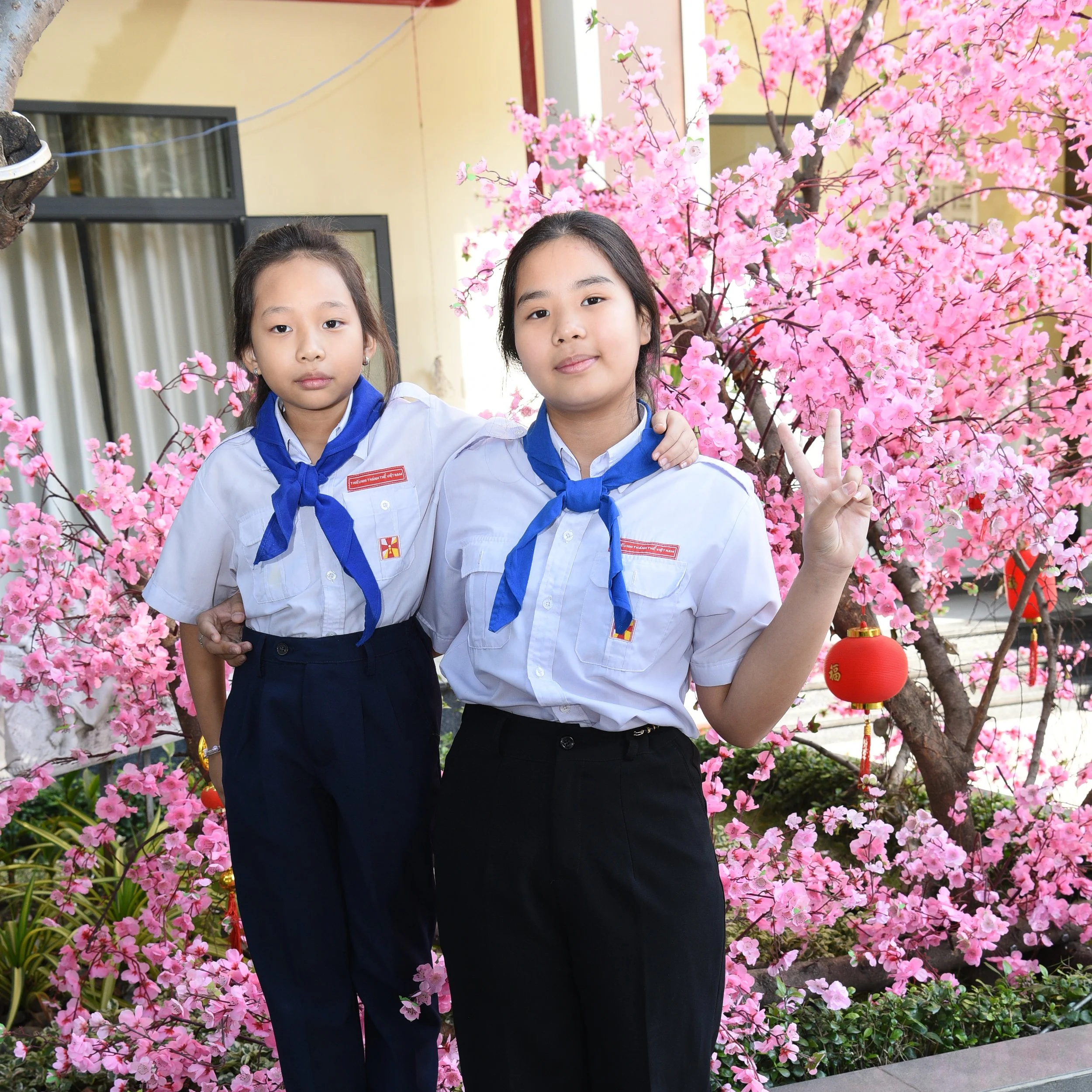 Two young girls in school uniforms posing in front of a pink flowering tree, with one girl making a peace sign.