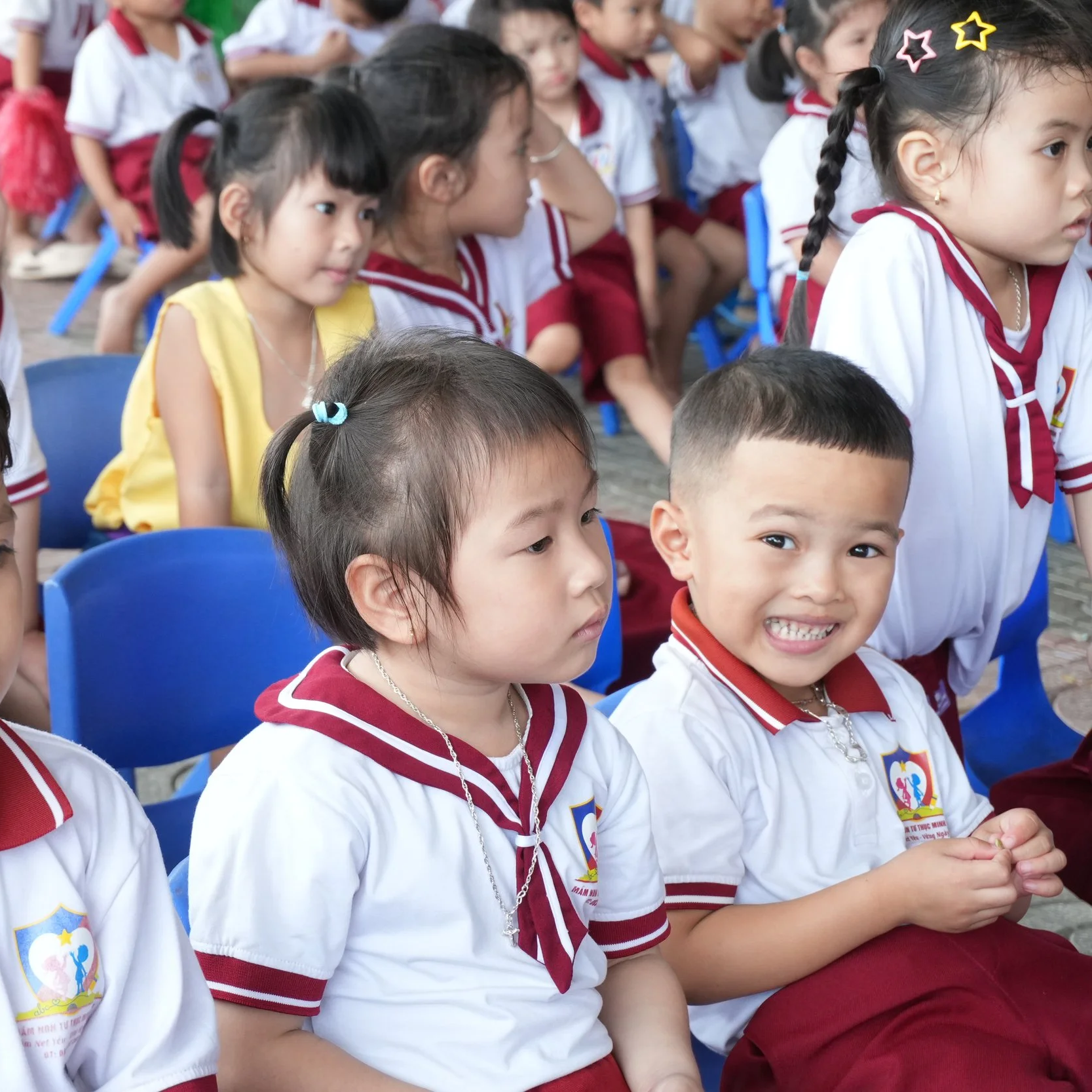 A group of young children sitting outdoors on blue chairs, wearing school uniforms with white shirts and maroon skirts or pants, with some children smiling and others looking serious.