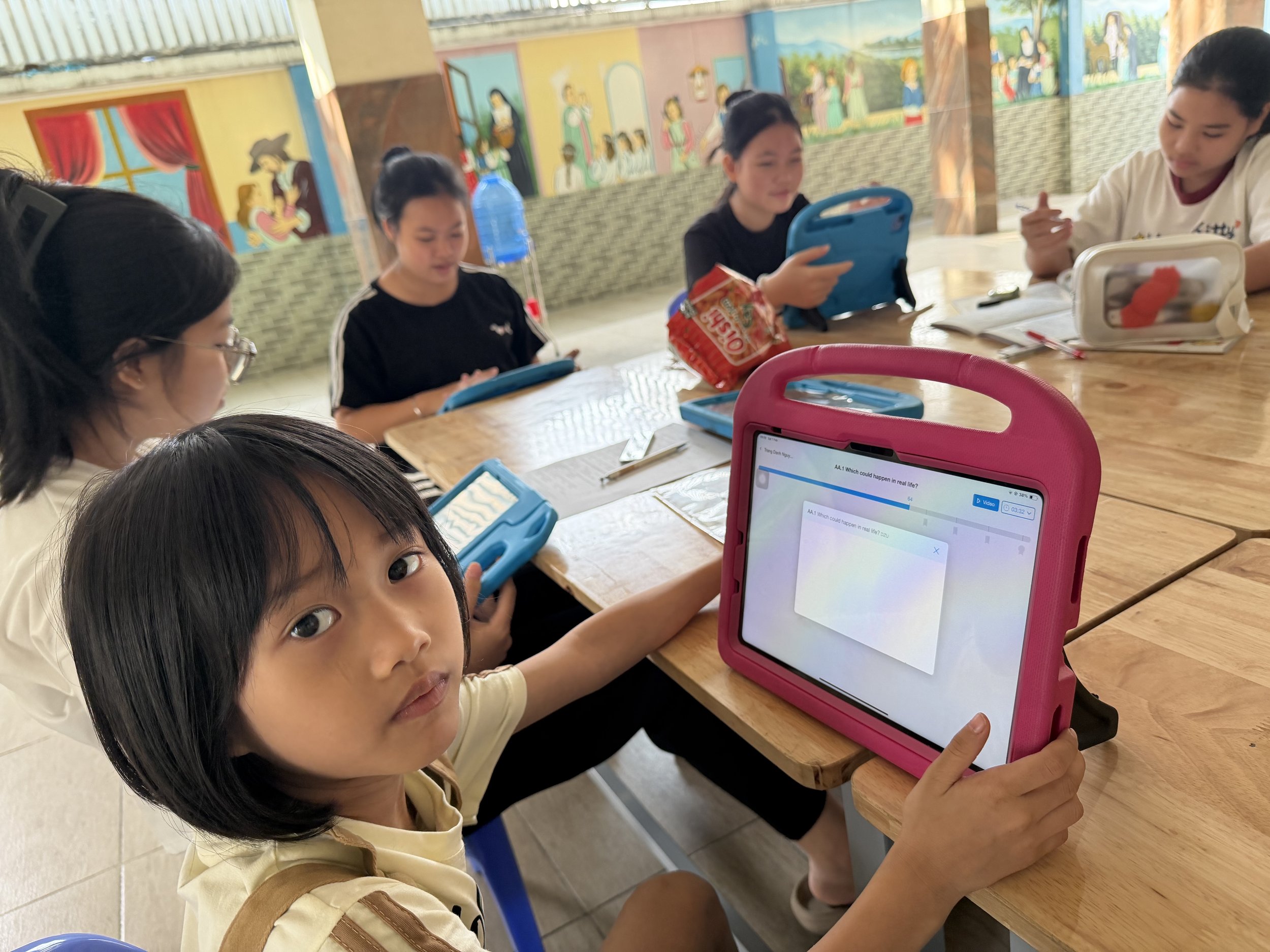 Young girl looking at the camera while sitting at a table with other children using tablets in a classroom that has colorful murals on the walls.