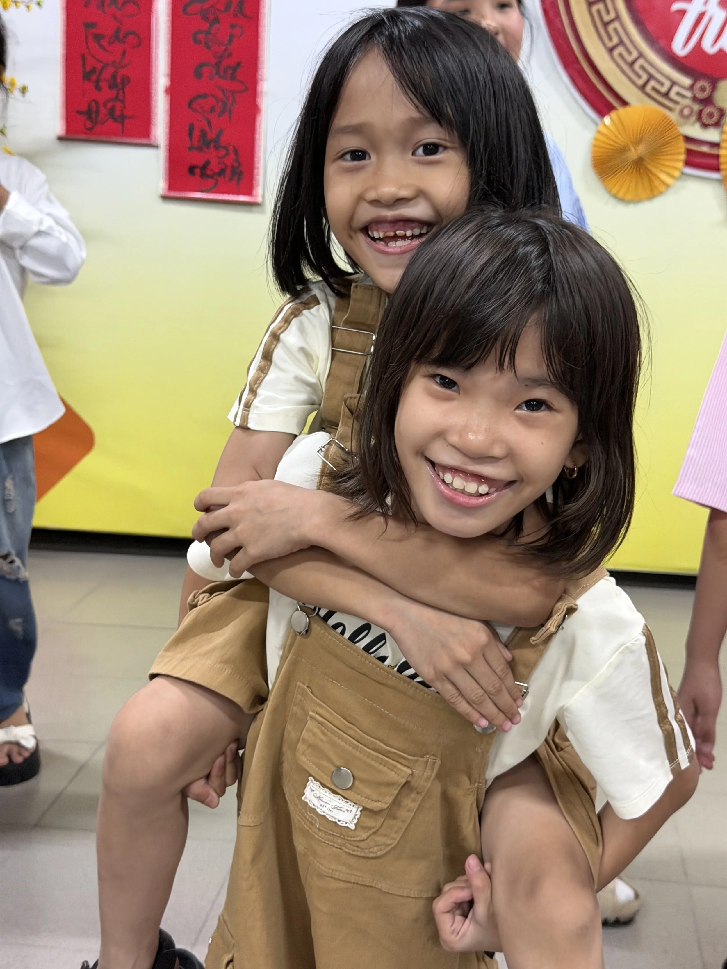 Two young girls with dark hair smiling, one giving the other a piggyback ride, with decorative red and gold banners and a yellow background.