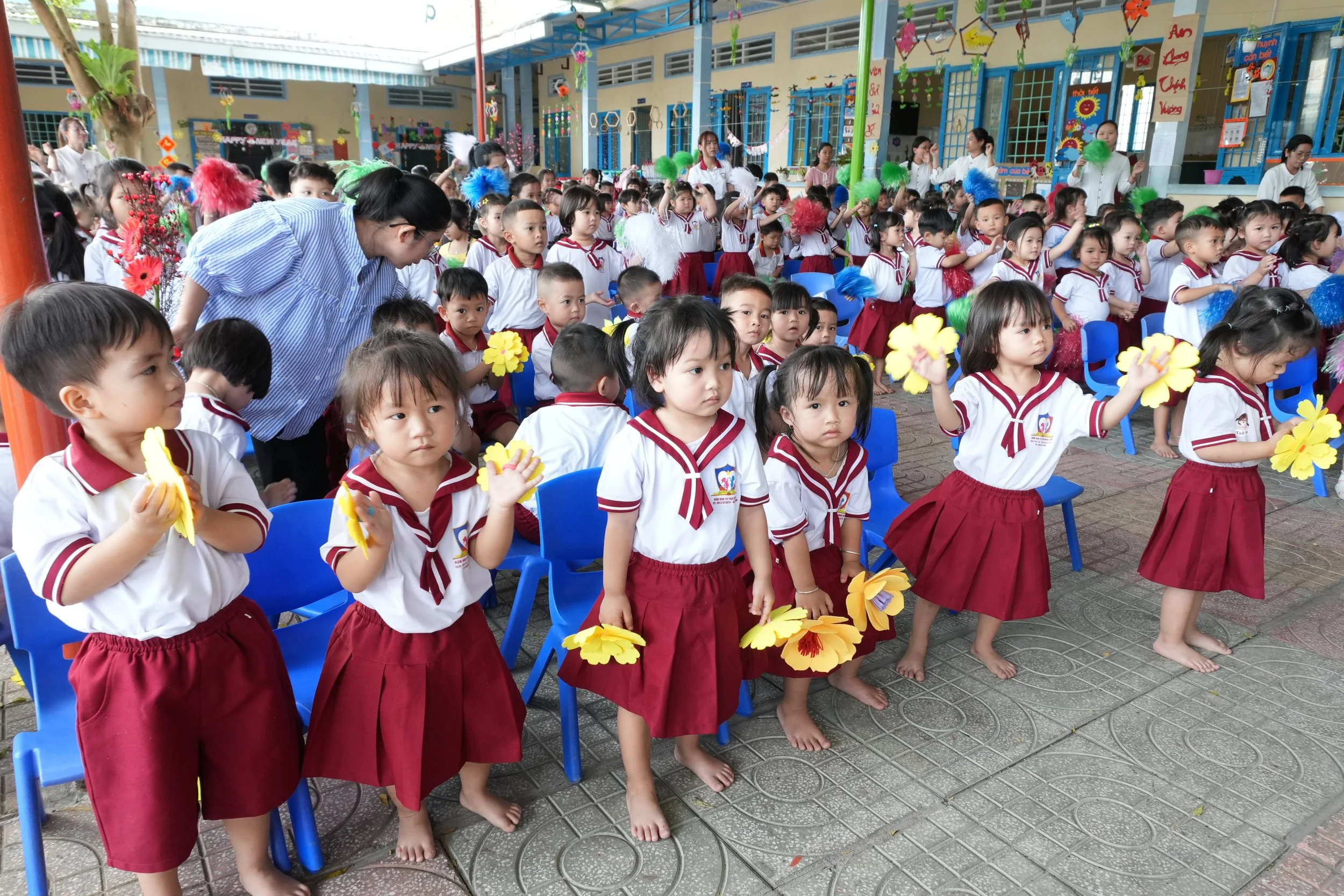 A group of young children in school uniforms, some barefoot, holding yellow craft flowers, participating in a school event outdoors with decorations and teachers around.