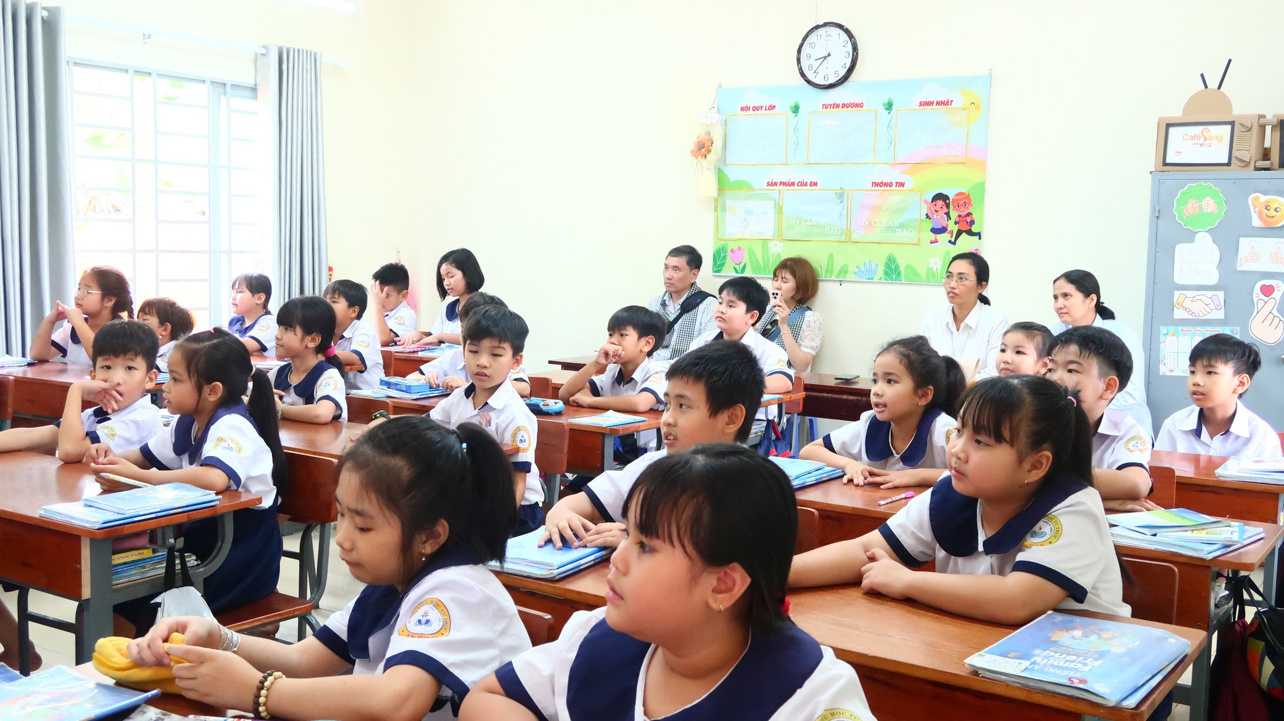 A classroom filled with young students wearing white and navy uniforms, sitting at wooden desks, listening attentively during a lesson. Several teachers and adults are seated at the back. The room has light-colored walls, a large window with curtains, and educational posters on the wall.