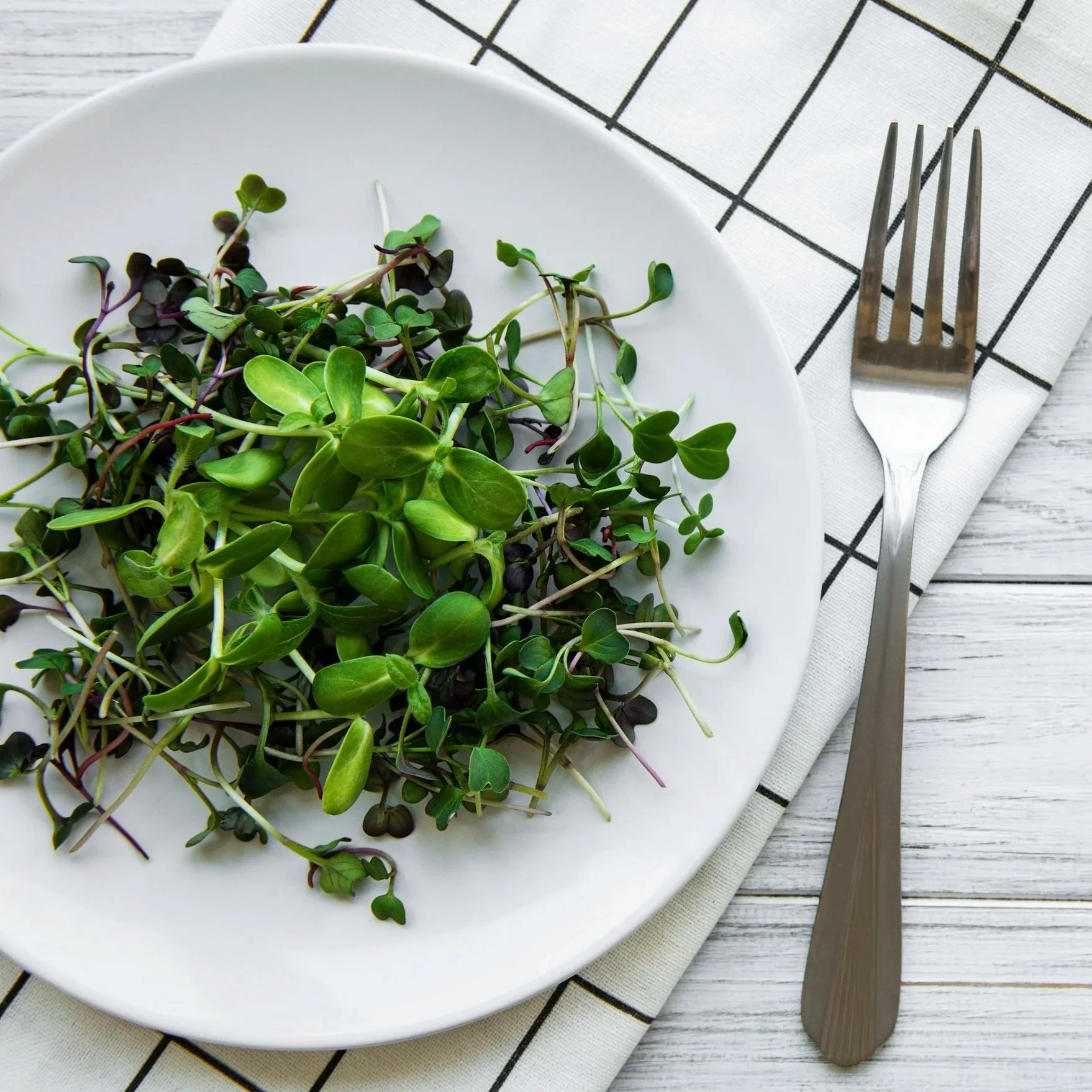 White bowl filled with various microgreens on a white plate, placed on a striped white cloth on a wooden table with a fork and knife to the side.