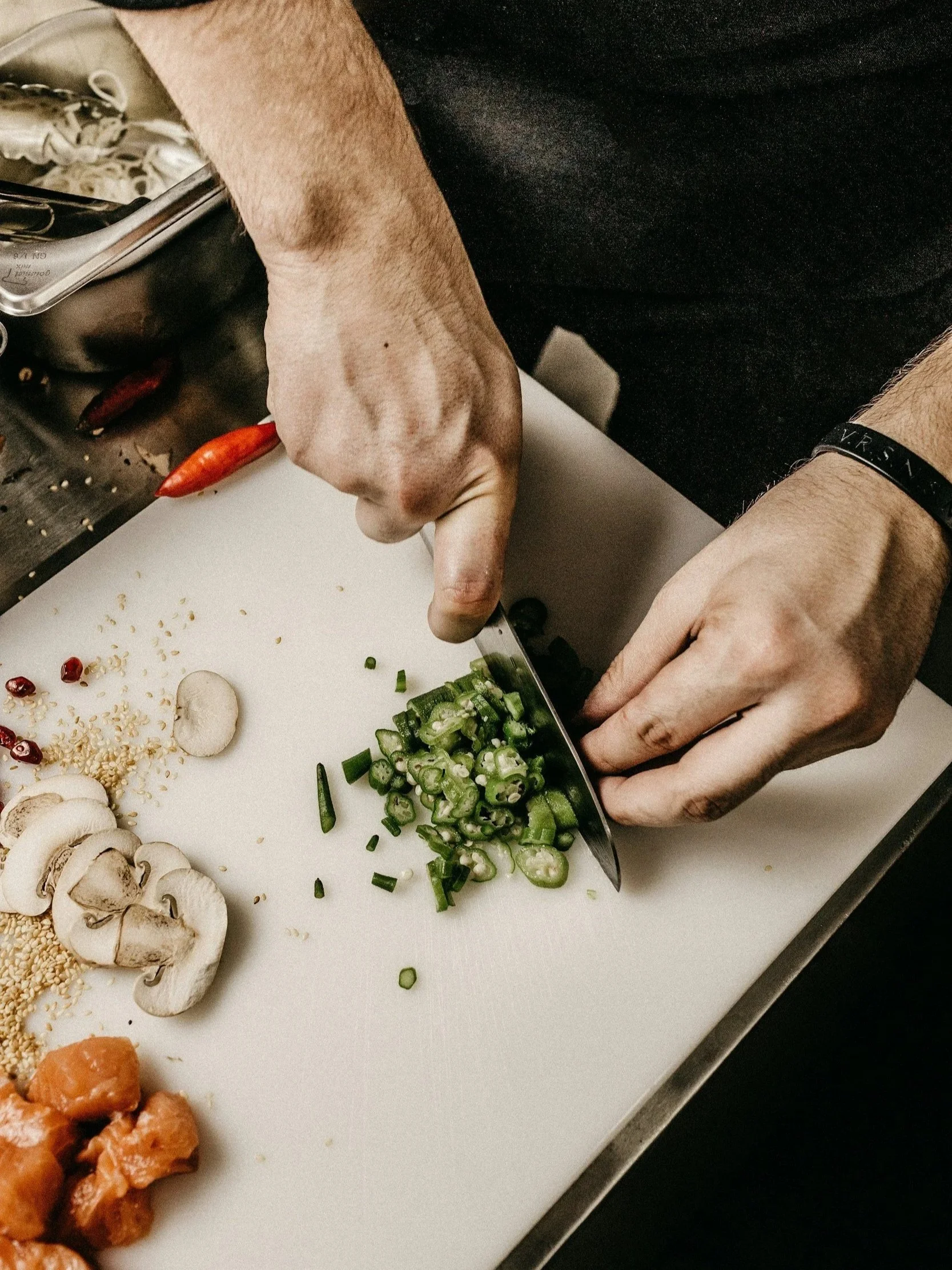 A person chopping green onions on a white cutting board, with mushrooms, sesame seeds, and other vegetables arranged nearby. For Chef inquiries.