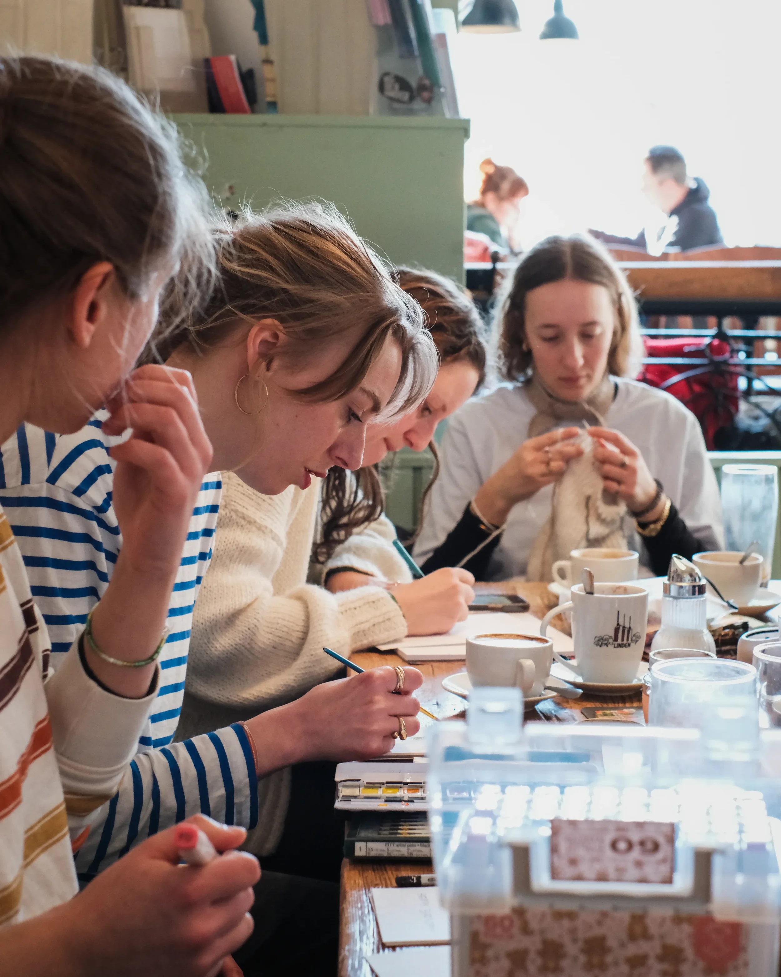 Fünf junge Erwachsene sitzen an einem Holztisch in einem Café und konzentrieren sich auf kreative Handarbeiten wie Zeichnen und Stricken. Auf dem Tisch stehen Kaffeetassen, Gläser und Bastelmaterialien in einer gemütlichen, hellen Atmosphäre