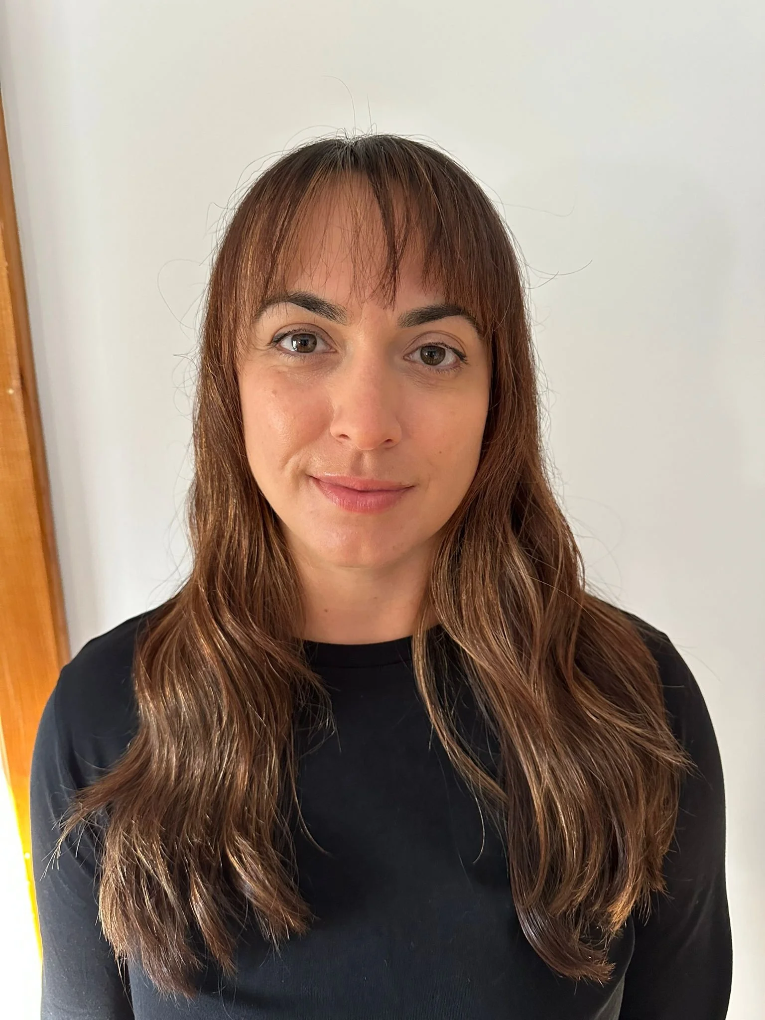 Portrait of a young woman with brown wavy hair and bangs, wearing a black top, standing in front of a plain light-colored wall.