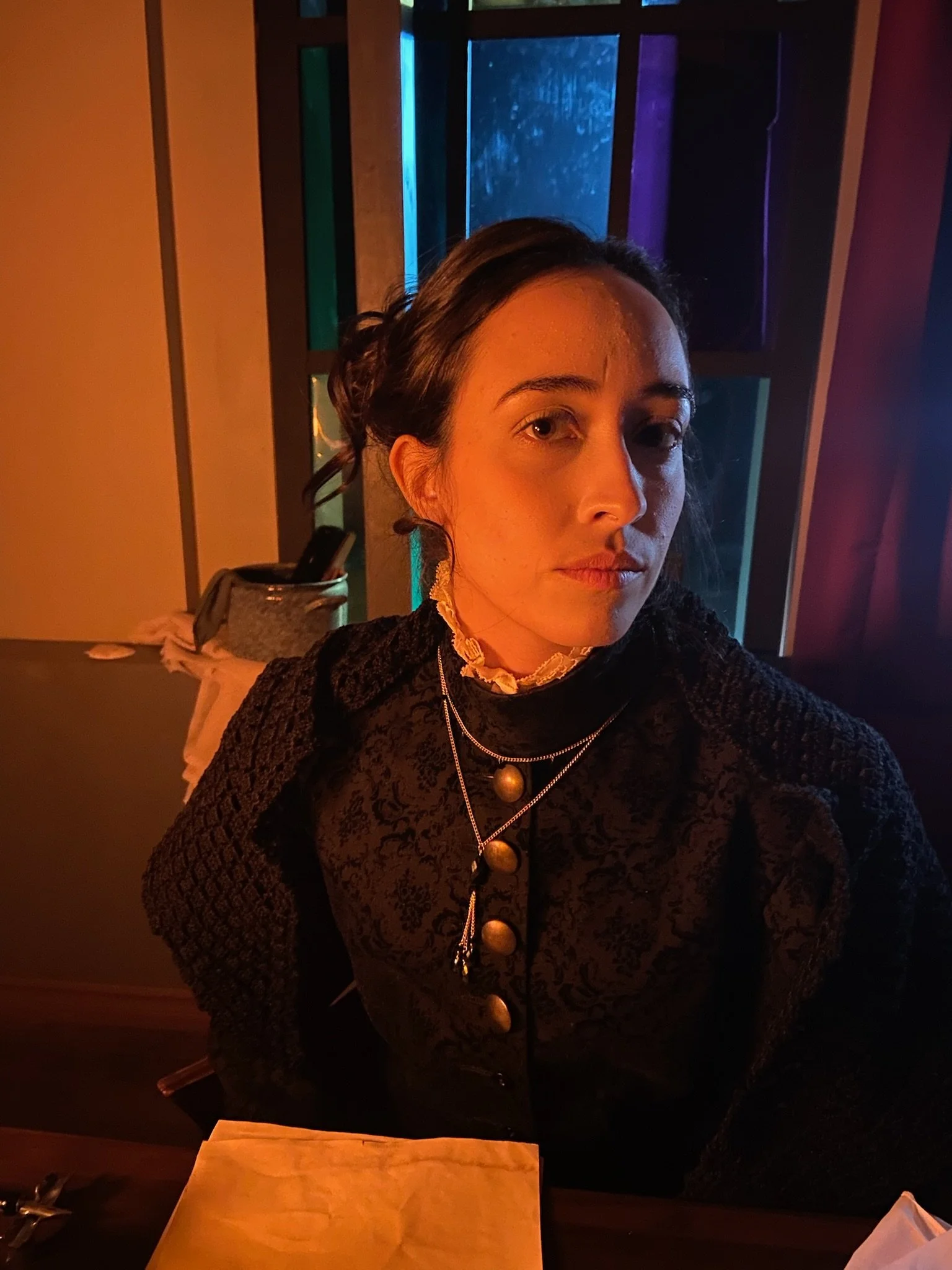 A woman with dark hair styled in a loose updo wearing a black textured jacket and layered necklaces with gold and silver beads, sitting at a table in a dimly lit indoor setting with colorful blue, purple, and orange lighting, a window with rain, and 