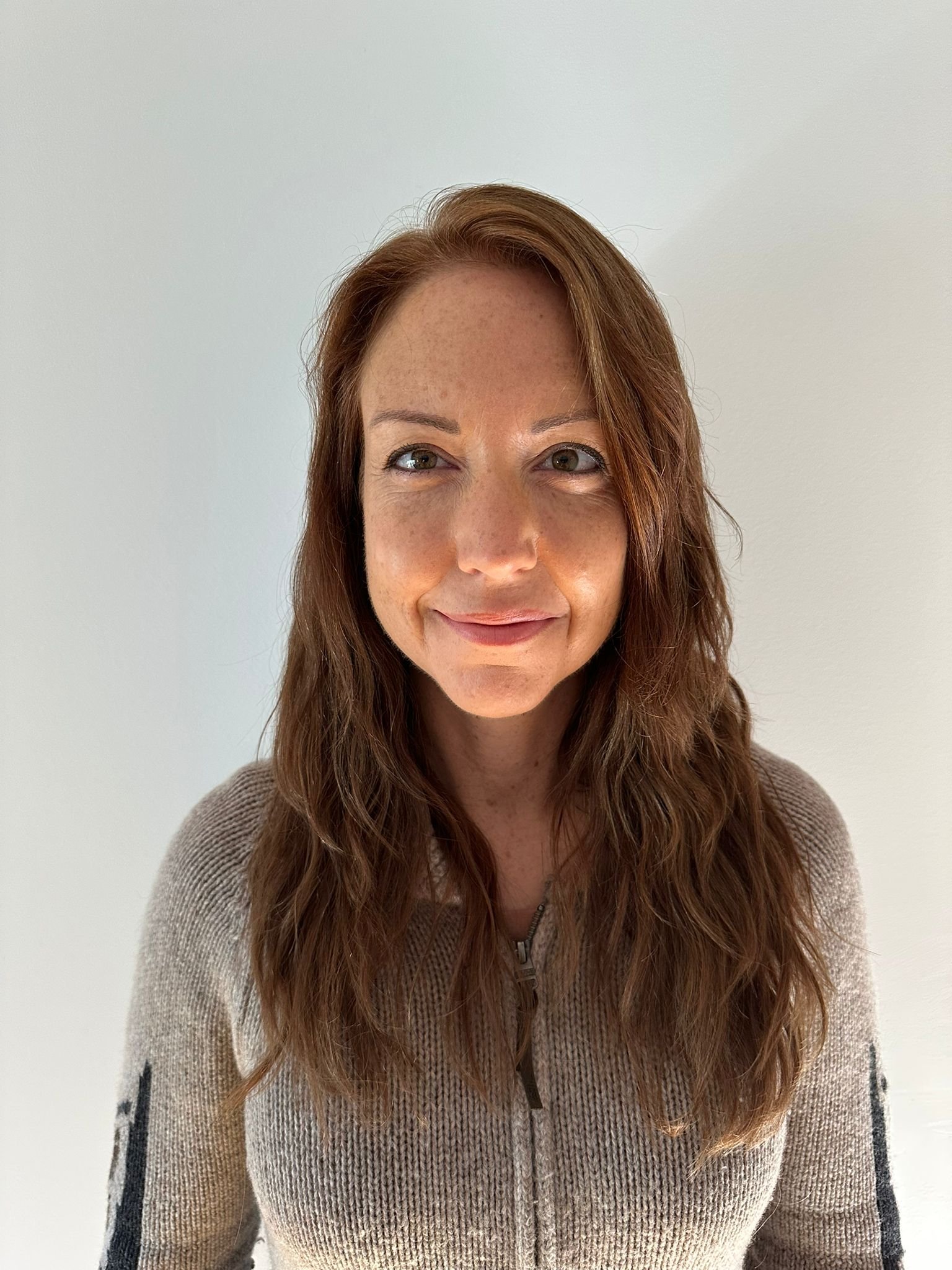 A woman with long, wavy brown hair and light skin, smiling gently, wearing a beige zip-up sweater, standing against a plain white wall.
