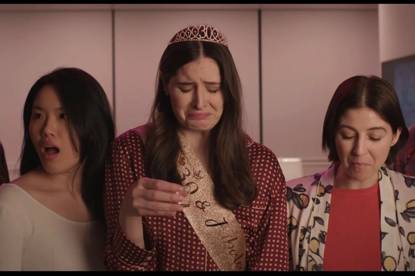 Three women at a celebration, one wearing a tiara and a sash, with emotional expressions.