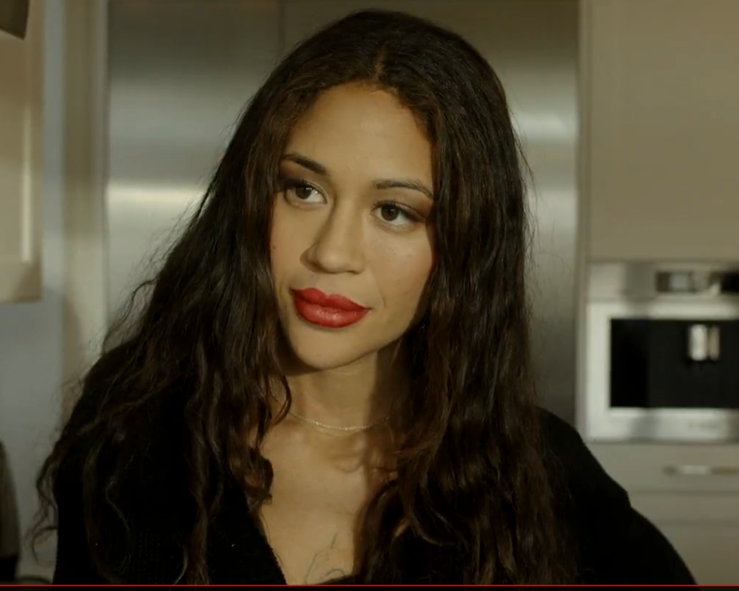Close-up of a woman with long, wavy brown hair, red lipstick, and wearing a black top, in a kitchen setting.