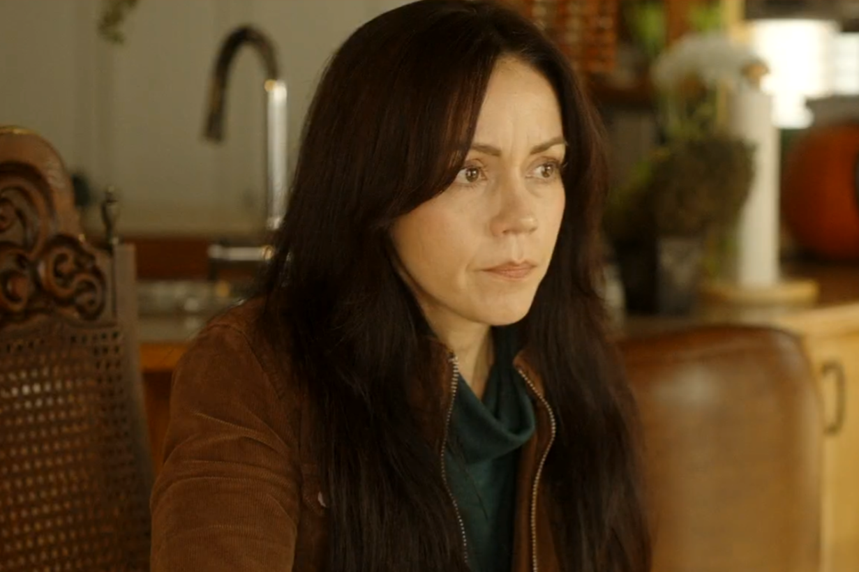 A woman with long dark hair and light skin, wearing a brown jacket, sitting at a table in a kitchen or dining area.