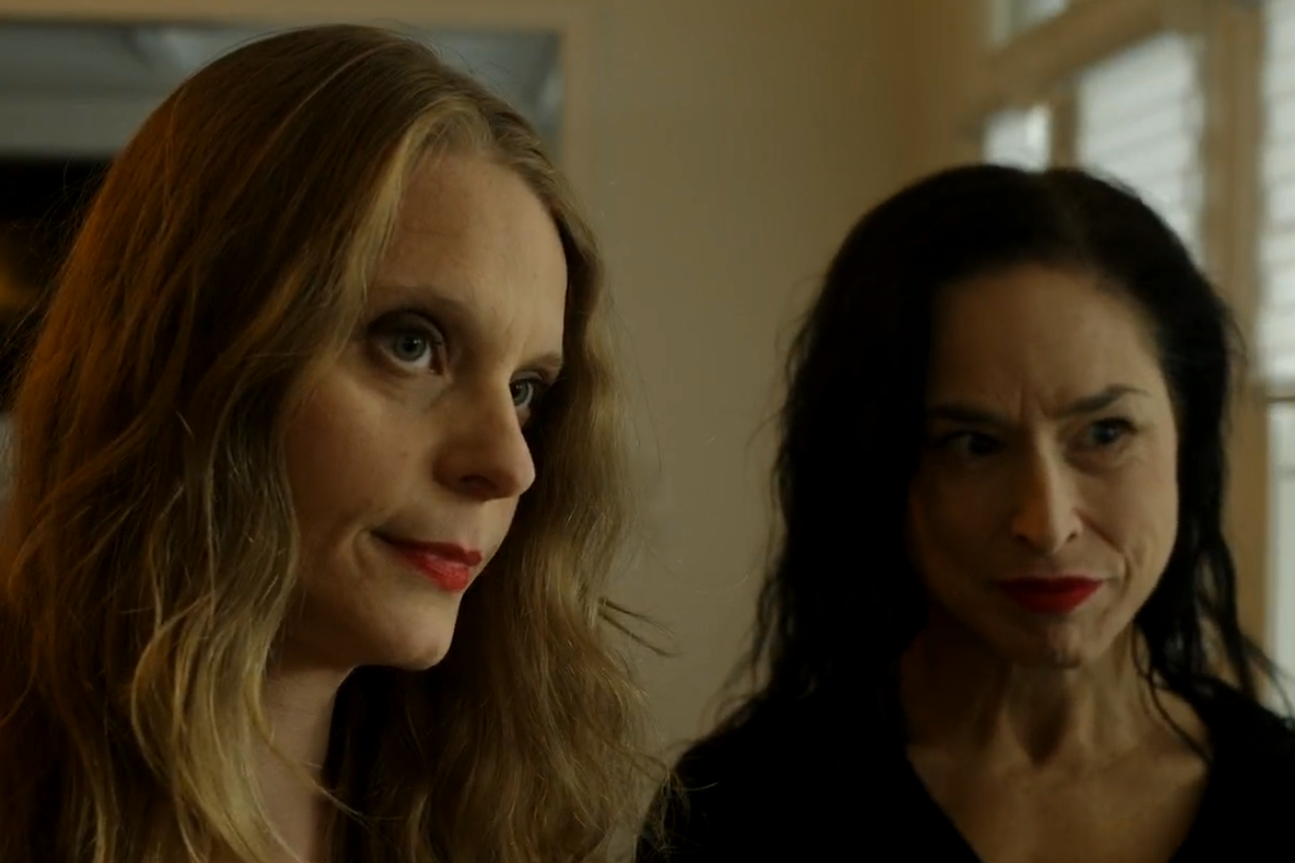 Two women with serious expressions, standing indoors near windows.