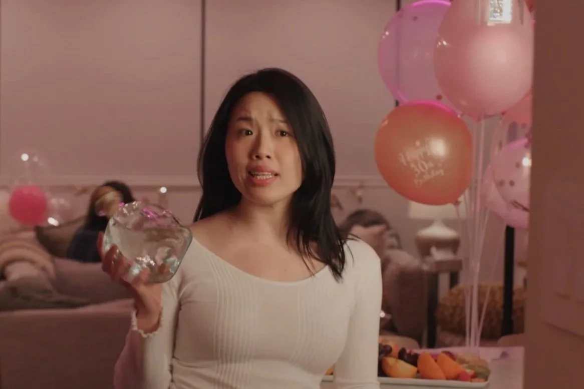 A woman with black hair wearing a white shirt holding a glass bowl, looking surprised or concerned, at a party with pink and peach balloons and a table with snacks in the background.