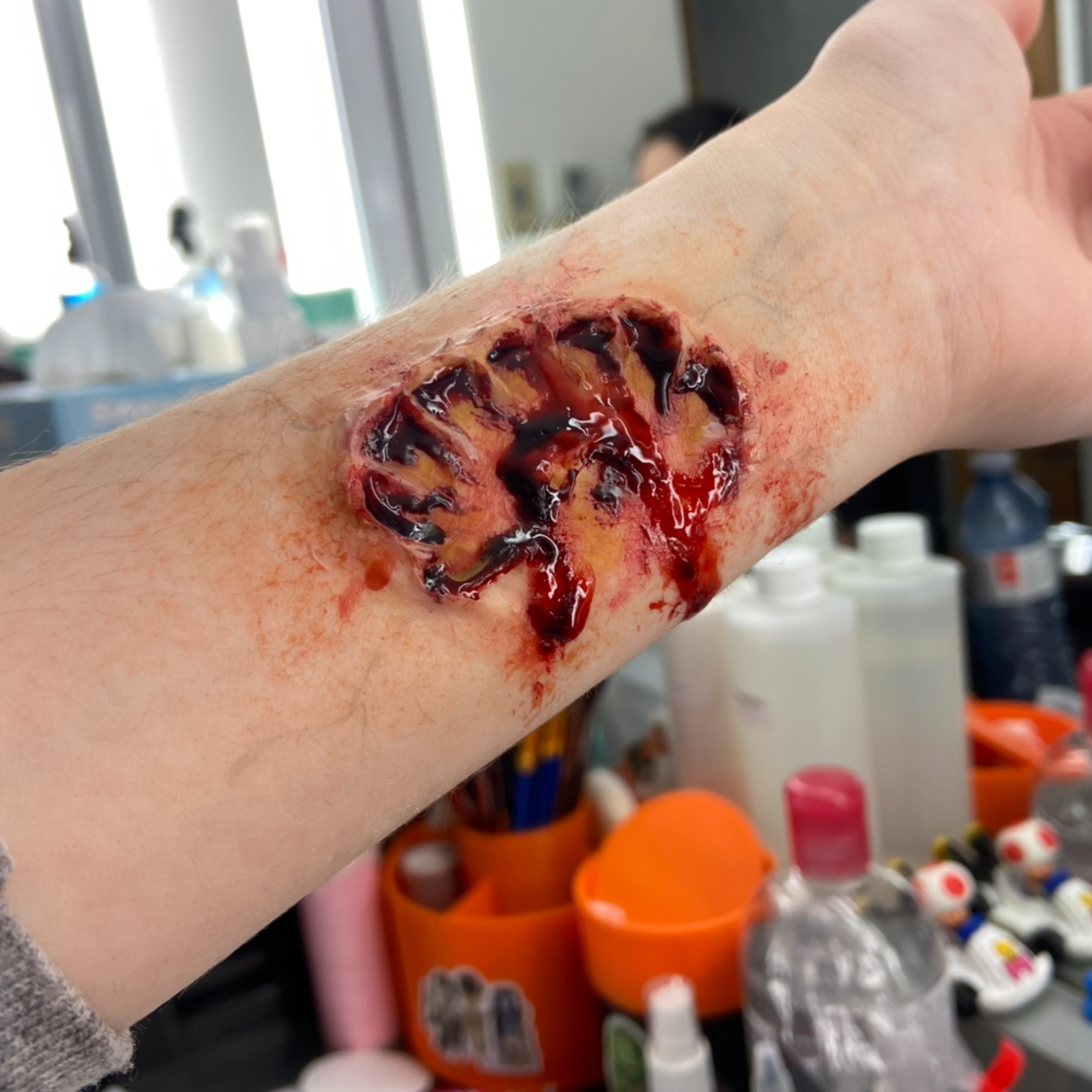A realistic prosthetic or special effects wound on the arm with exposed flesh, muscles, and blood, simulating a severe injury or burn.
