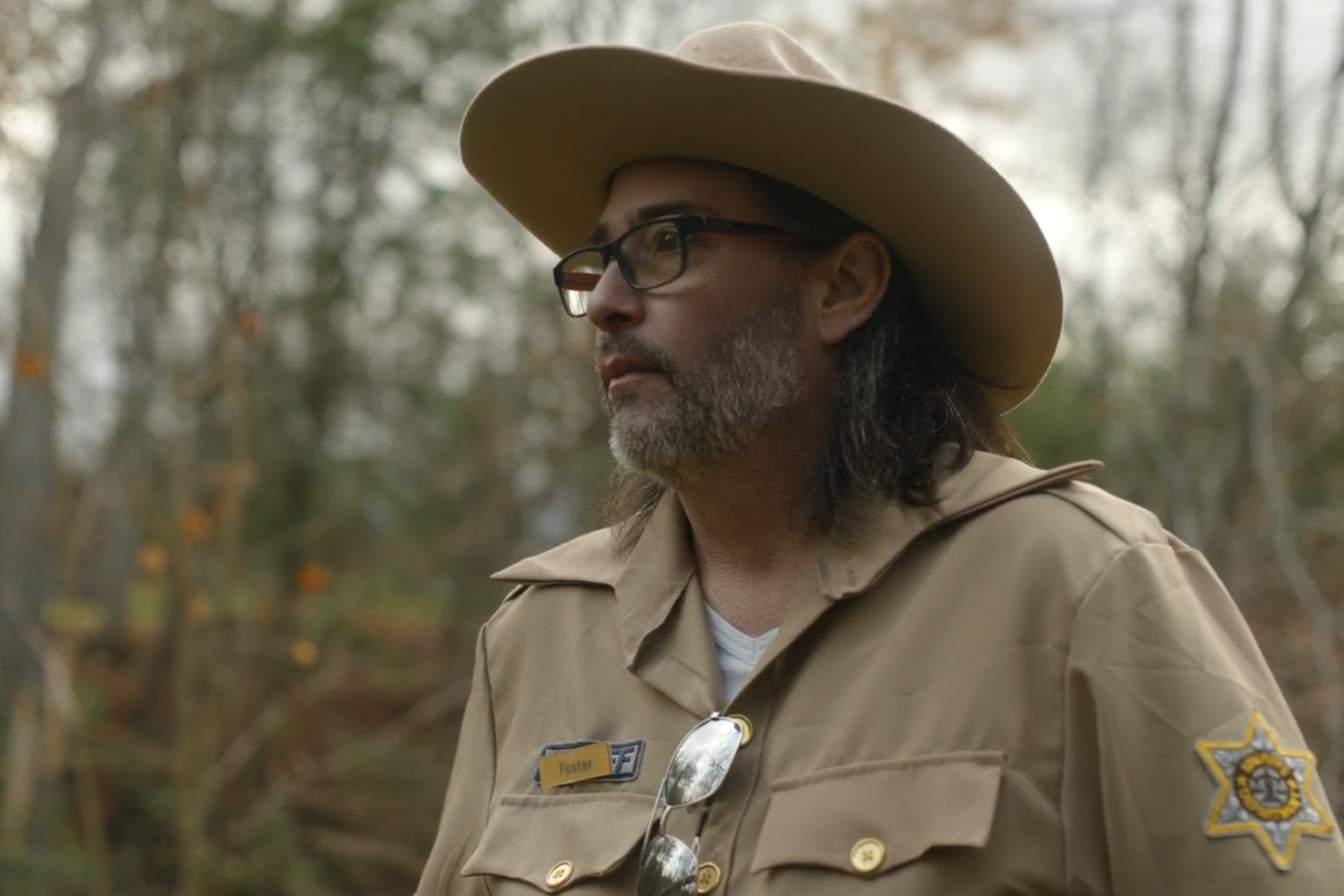 Man in a sheriff's uniform and glasses, wearing a wide-brimmed hat, standing outdoors in a wooded area.