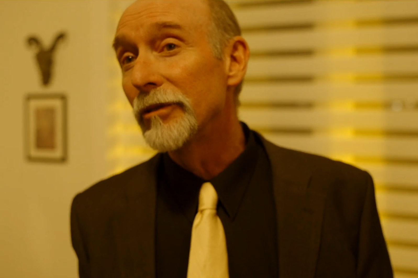 A man with a gray beard and bald head, wearing a black shirt, cream-colored tie, and a dark blazer, standing indoors against a yellow wall with framed pictures and a blurred pattern.
