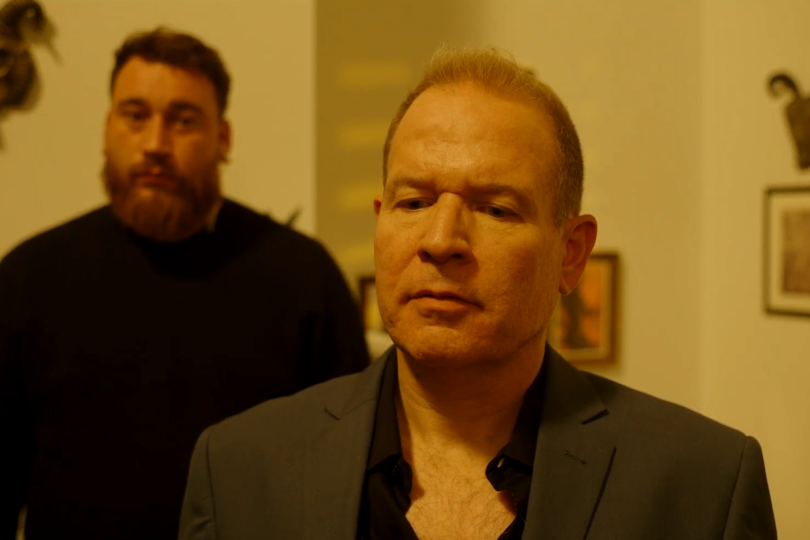 Two men in a room with framed pictures on the wall; one man in the foreground wearing a dark blazer with a serious expression, the other man in the background wearing a black shirt with a beard.