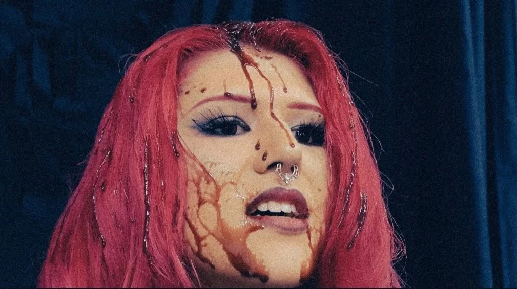 A woman with pink hair, makeup, and facial piercings, with a dark background.