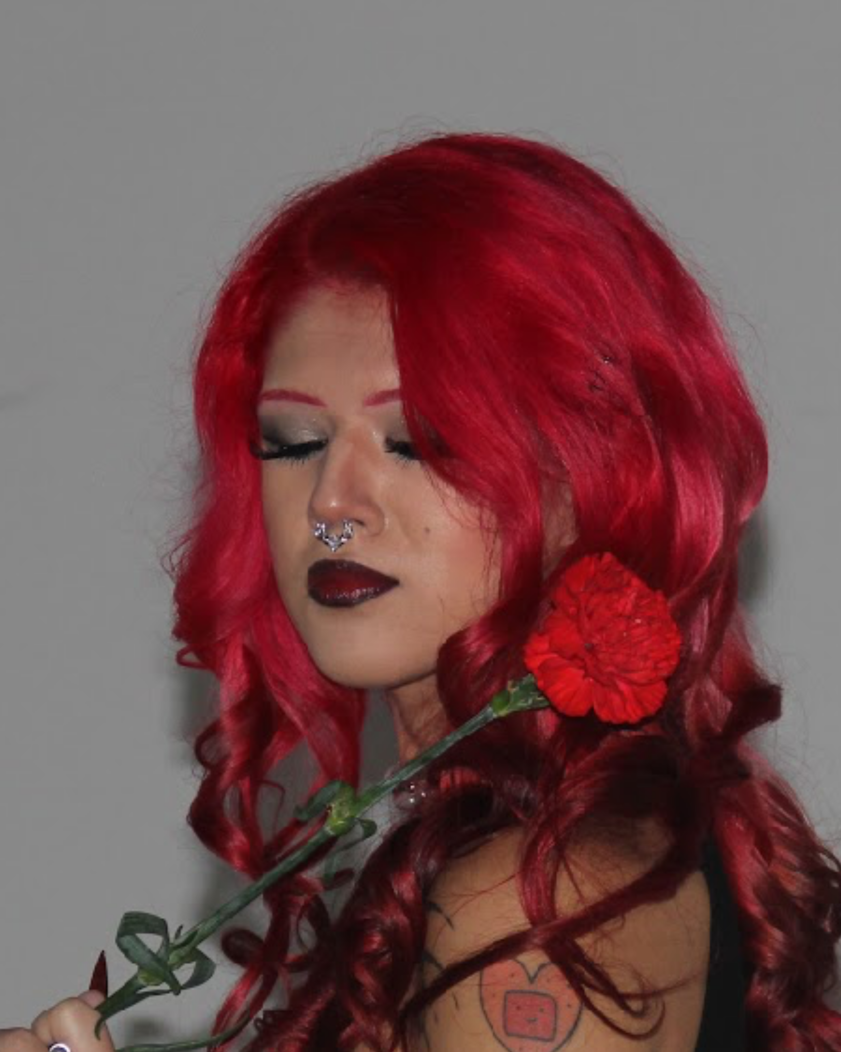 Woman with bright red, curly hair holding a red carnation flower near her face, with her eyes closed and dark makeup.