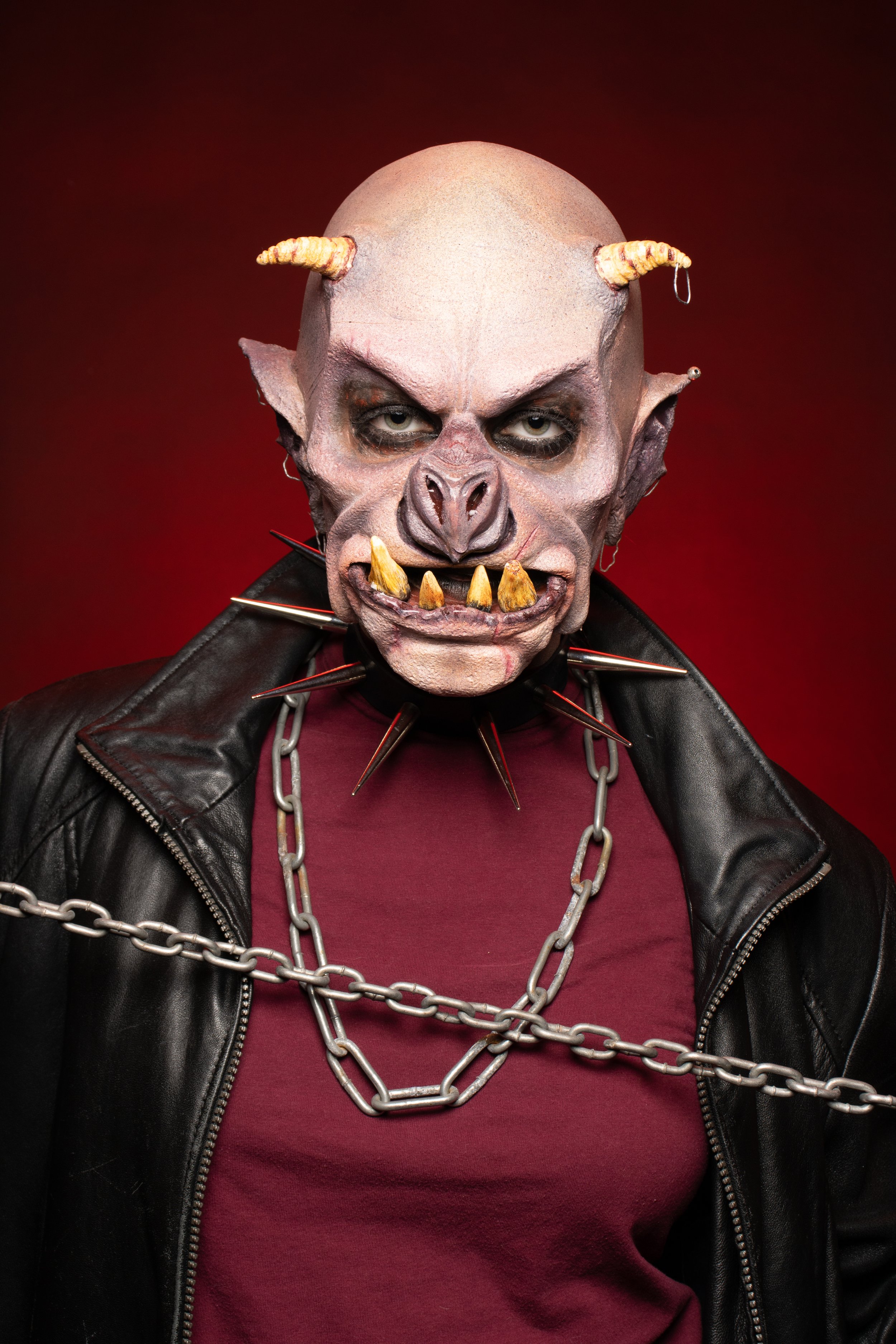 A person dressed as a scary, orc-like creature with a bald head, horns, and sharp teeth, wearing a black leather jacket, maroon shirt, and chains around the neck, against a red background.
