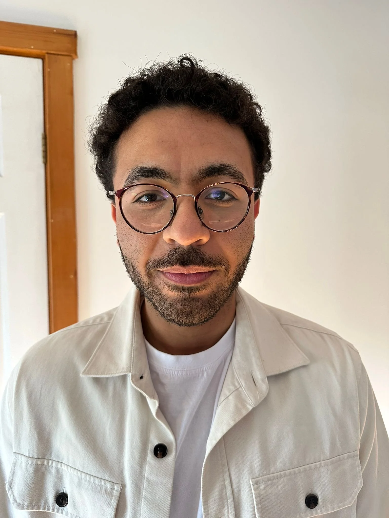 A young man wearing glasses and a white shirt standing indoors against a plain white wall.