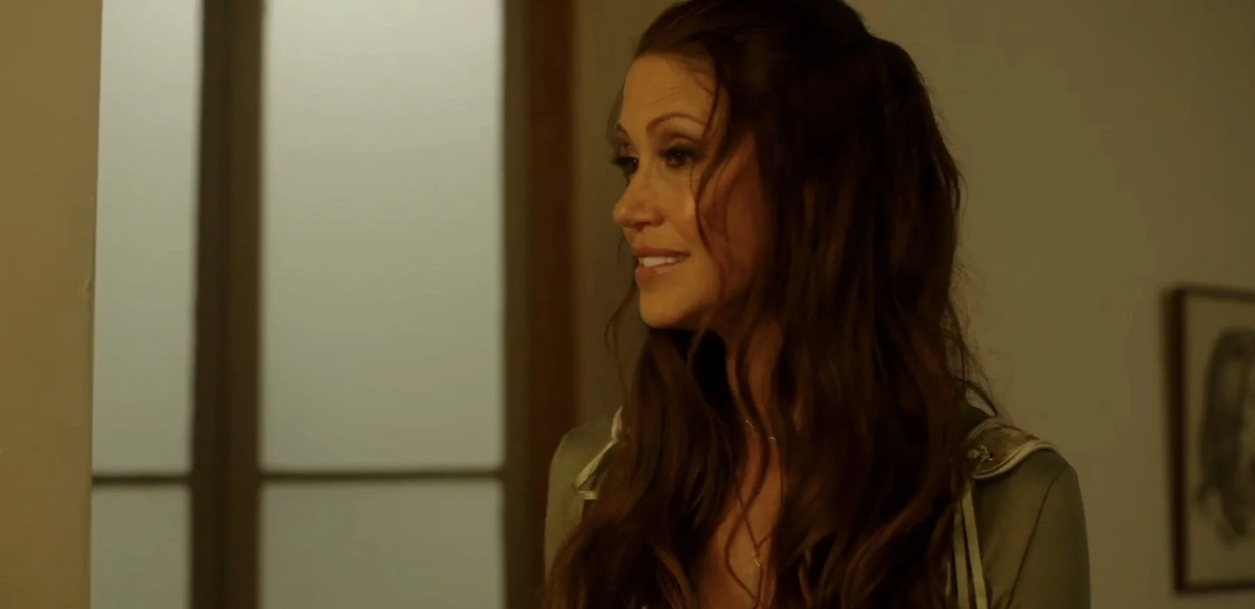 A woman with long, wavy brown hair smiles while looking to the side indoors.