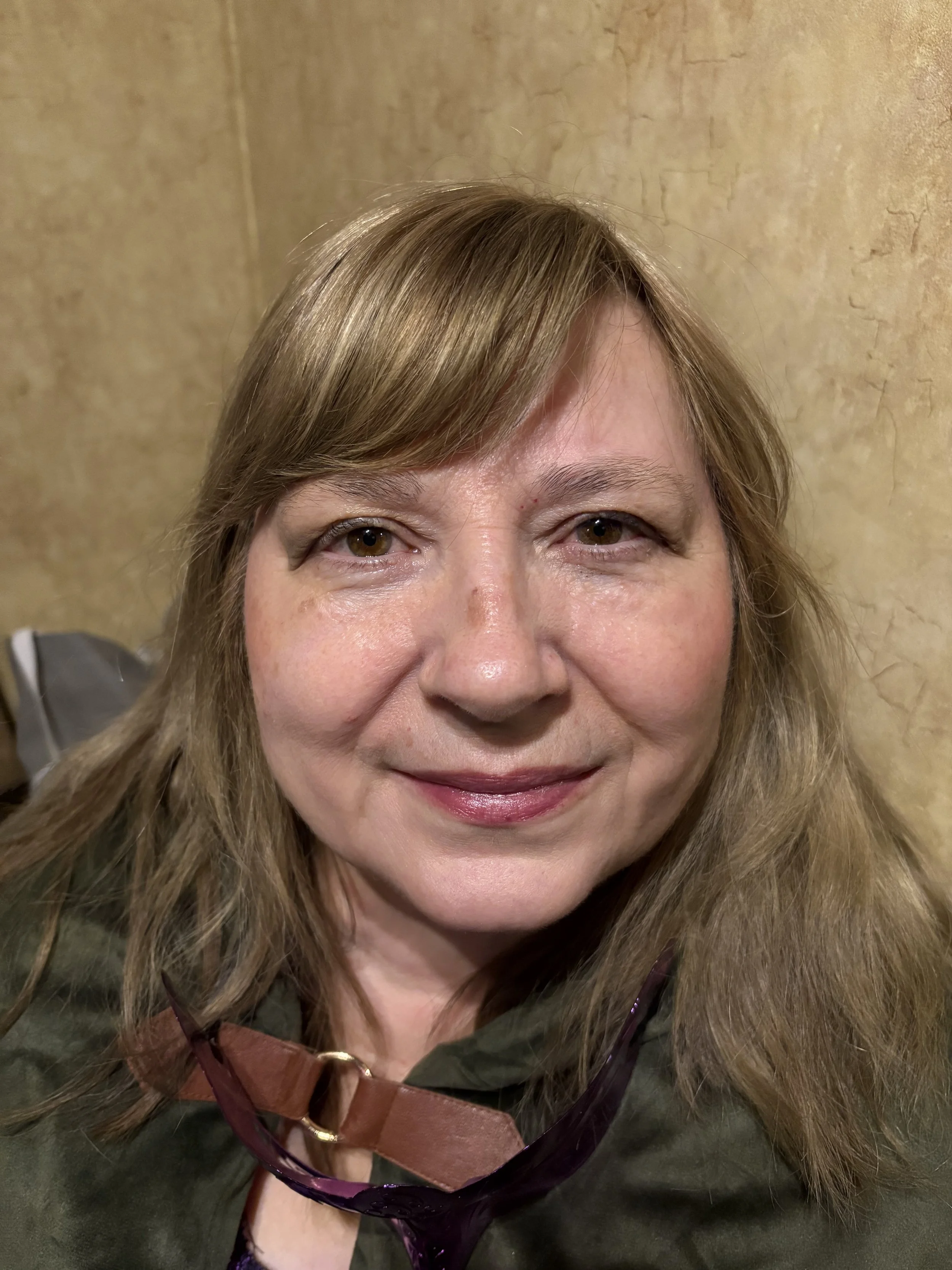 A woman with light brown hair and brown eyes smiling at the camera, wearing a green jacket with a brown strap and purple sunglasses hanging around her neck, with a textured beige wall background.