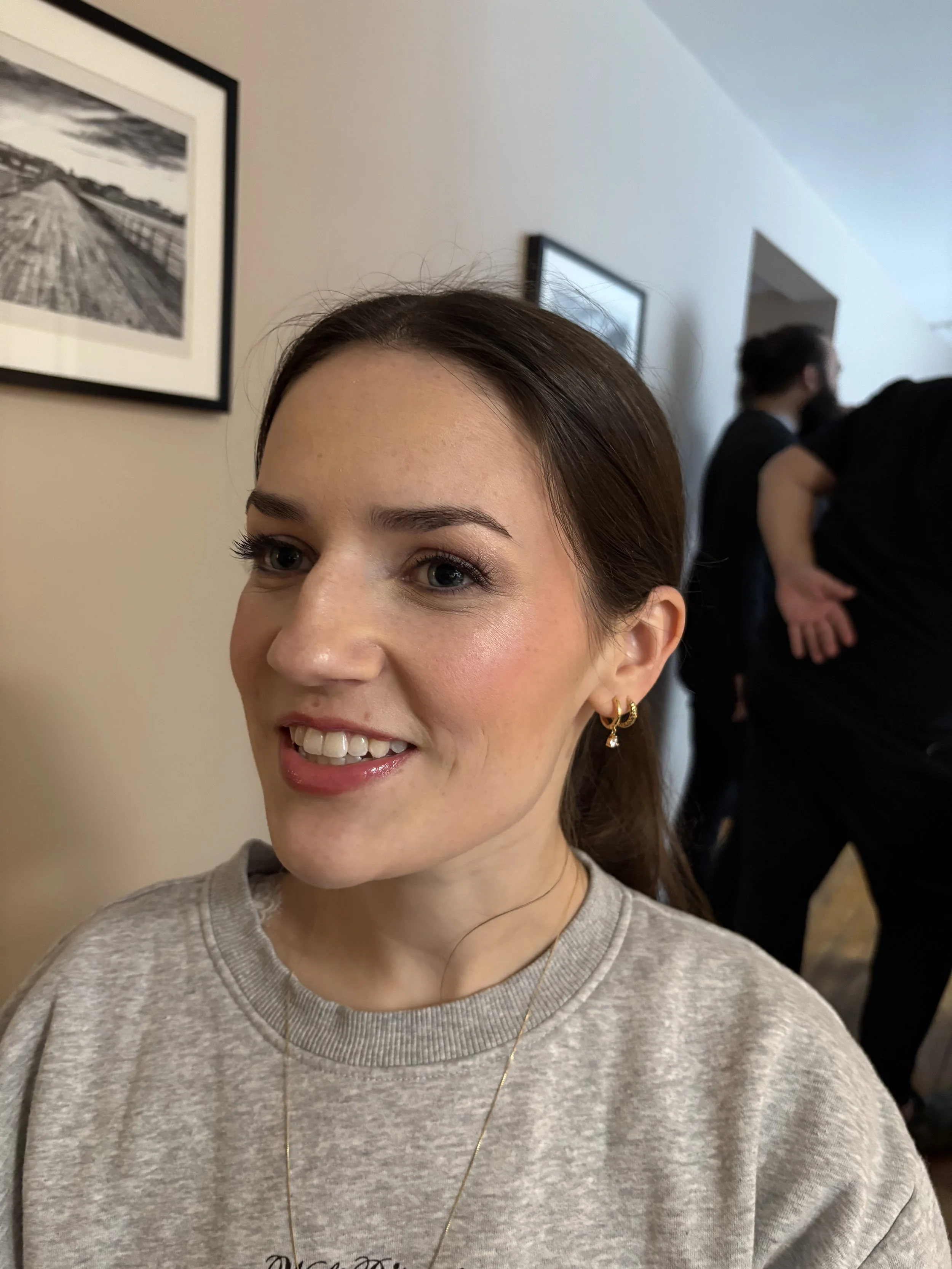A close-up of a woman with brown hair, wearing gold earrings and a gray sweatshirt, smiling inside a room with framed black-and-white pictures on the wall and people in the background.