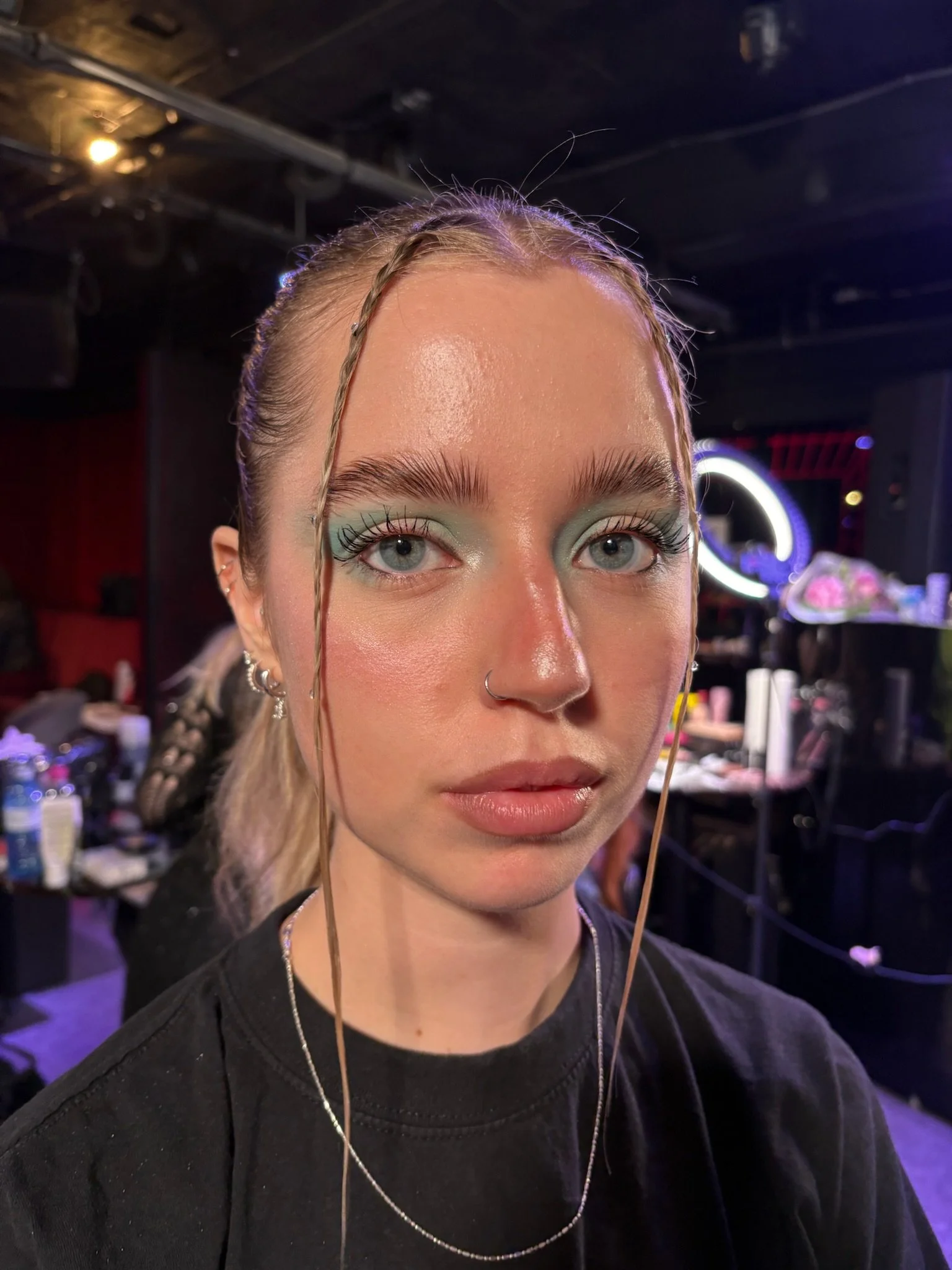 Close-up of a young woman with makeup, including pastel blue eyeshadow, thin eyebrows, and a nose ring, in a backstage or makeup studio setting with blurred background of makeup tools and people.