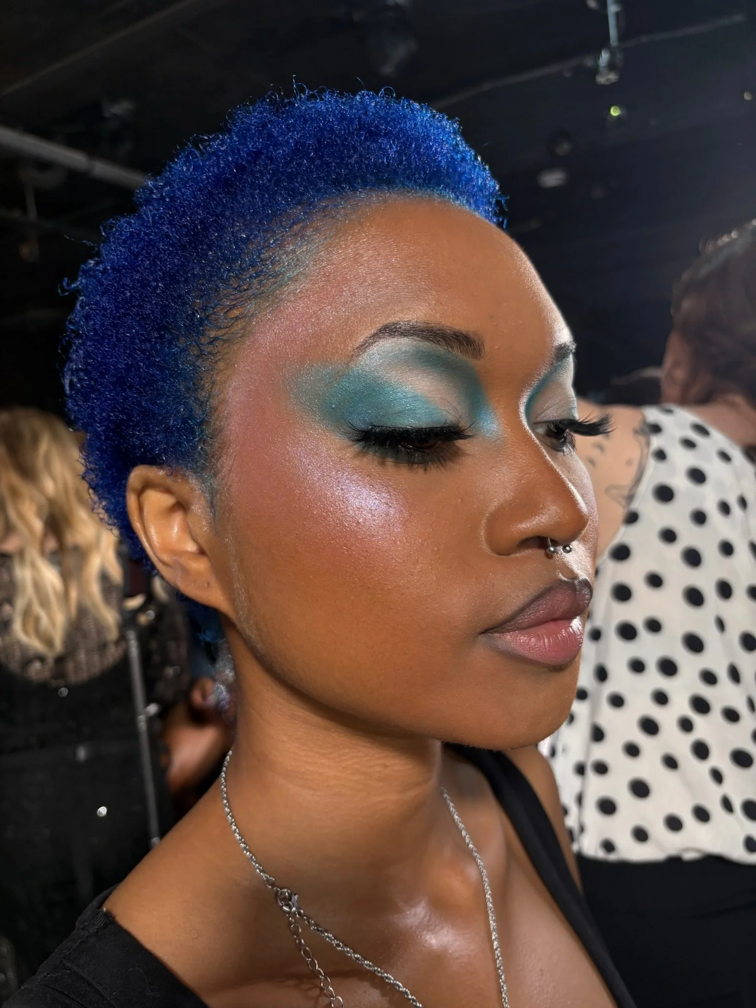 Close-up of a woman with vibrant blue hair, dramatic turquoise and silver eye makeup, long dark eyelashes, and a silver septum piercing, wearing a necklace and black top, in a crowded indoor setting.