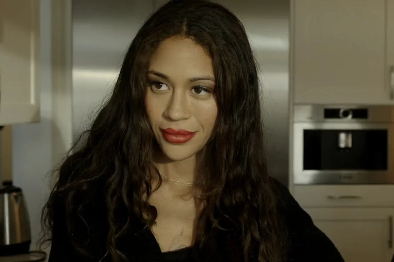 A woman with long, dark curly hair and red lipstick, looking to the side, standing in a kitchen.
