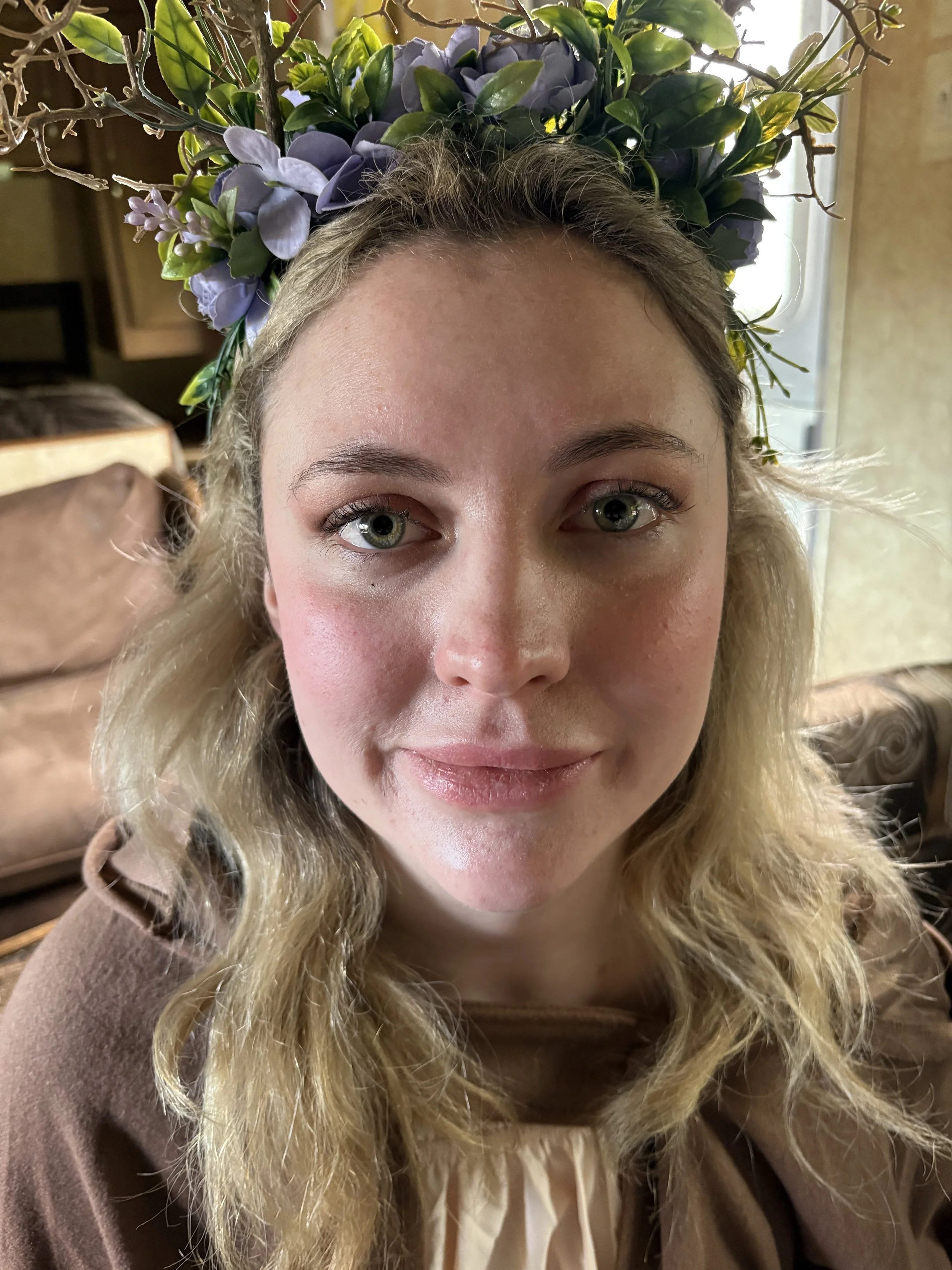 A woman with blonde hair and blue eyes wearing a floral crown of purple and green flowers, smiling at the camera in a cozy indoor setting.