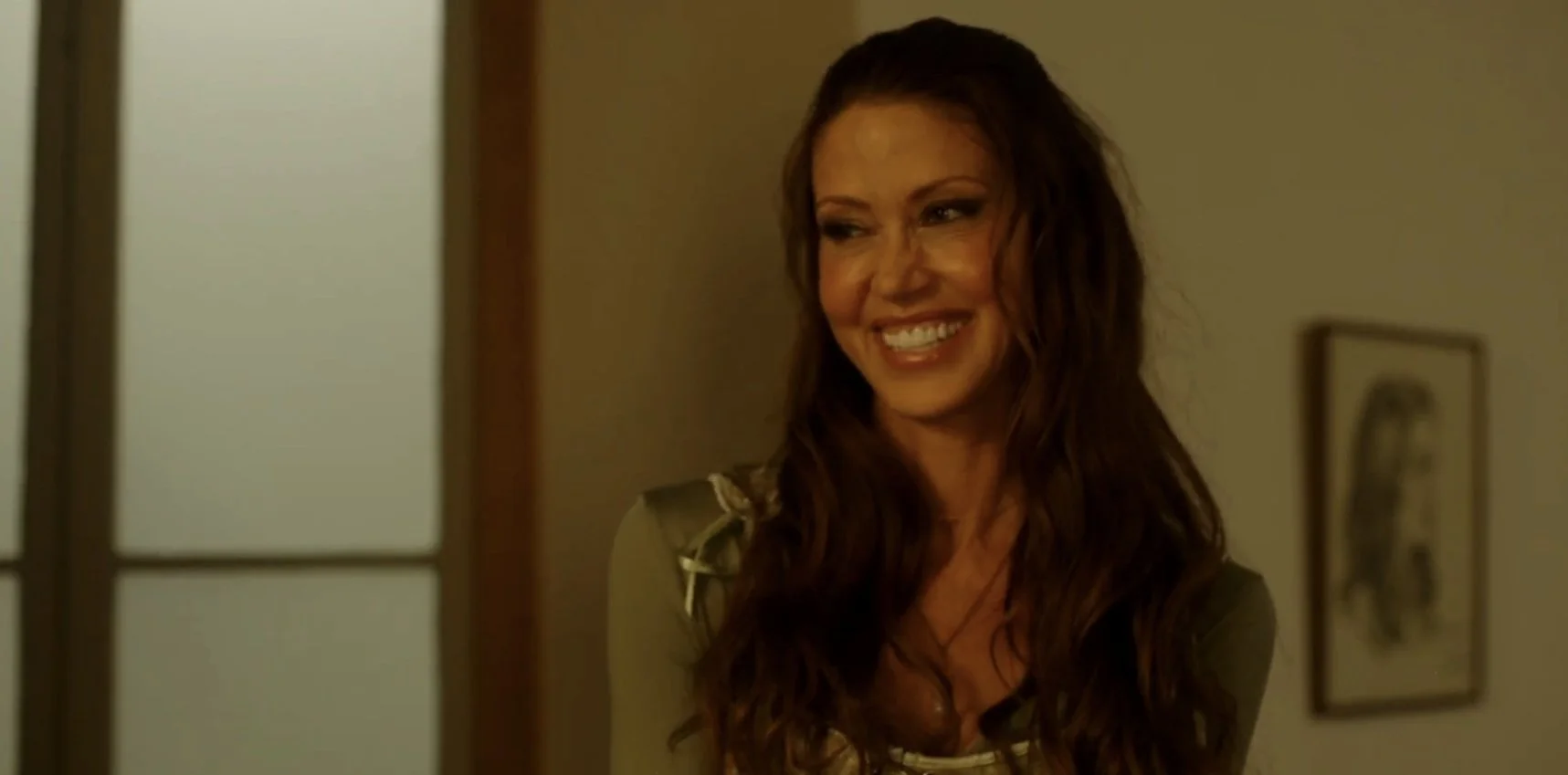 A woman with long, wavy brown hair is smiling, standing in a room with beige walls and a framed picture hanging in the background.