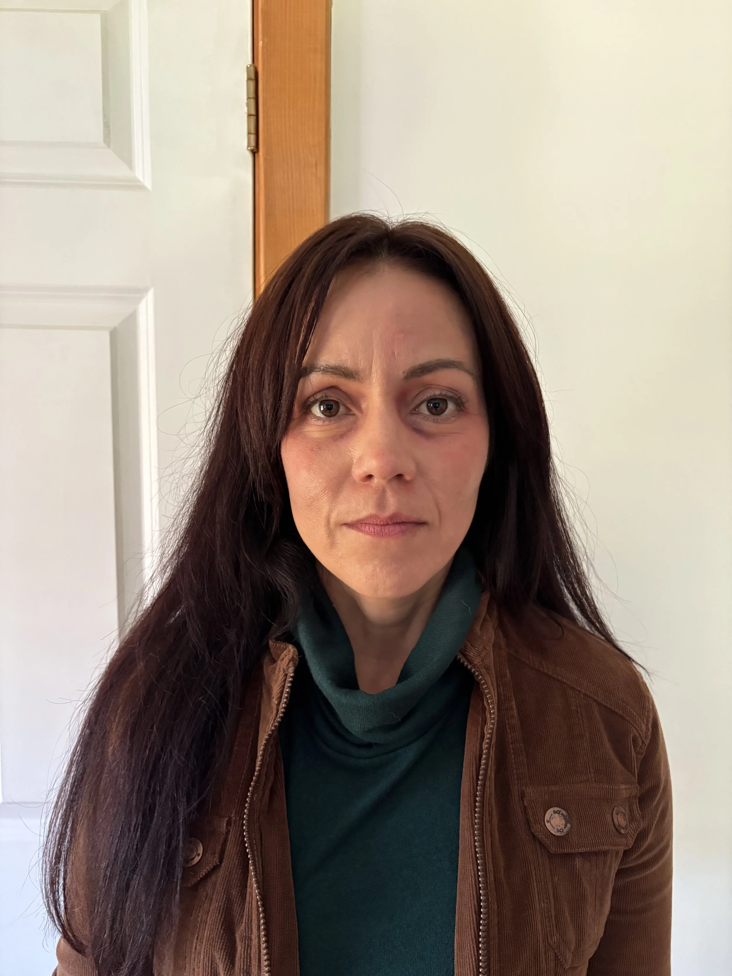 A woman with long dark hair and a serious expression, wearing a green top and a brown jacket, standing indoors with plain walls in the background.
