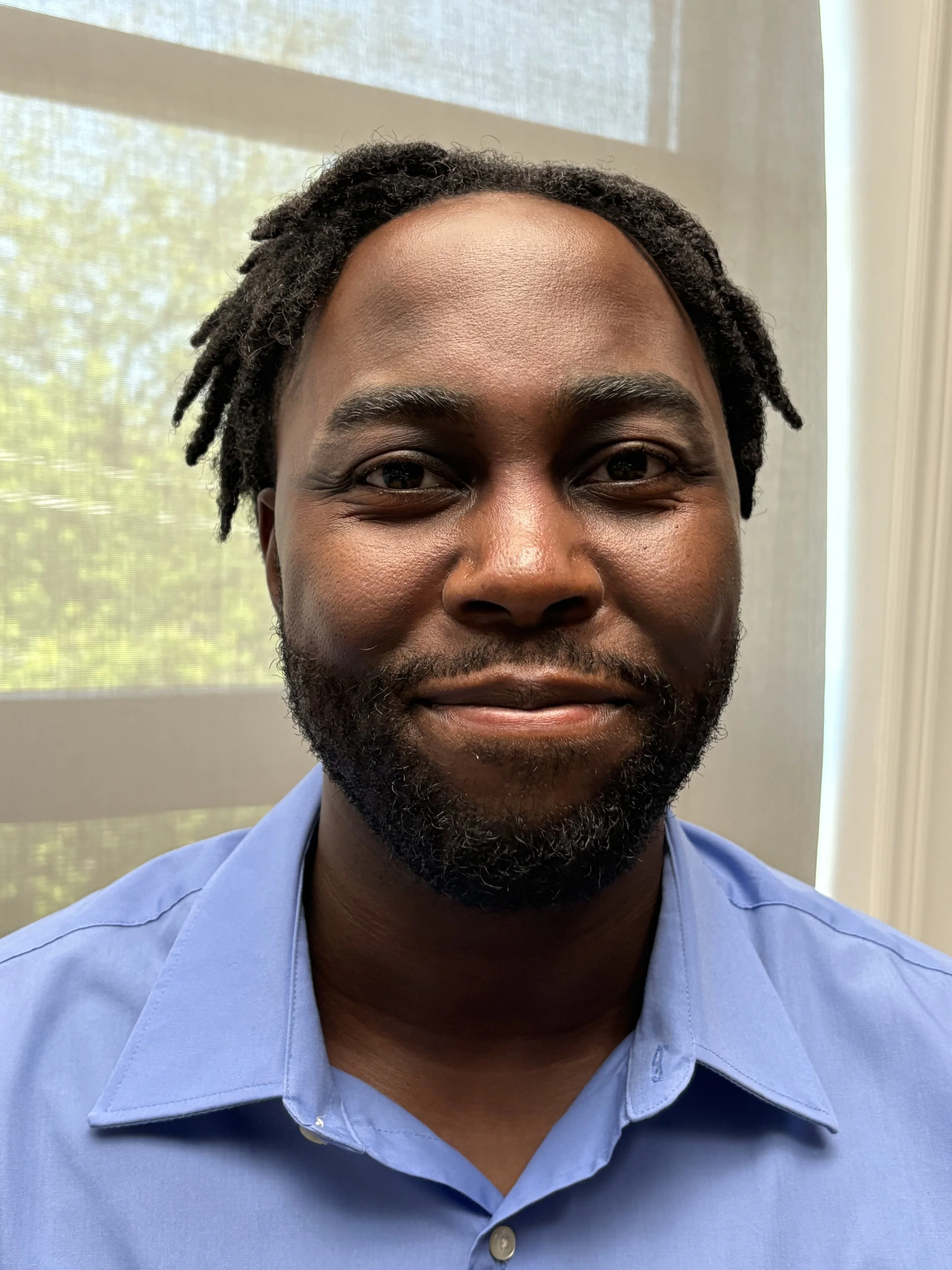 Close-up of a smiling man with dark skin, black beard, wearing a light blue collared shirt, standing in front of a window with a screen and greenery outside.