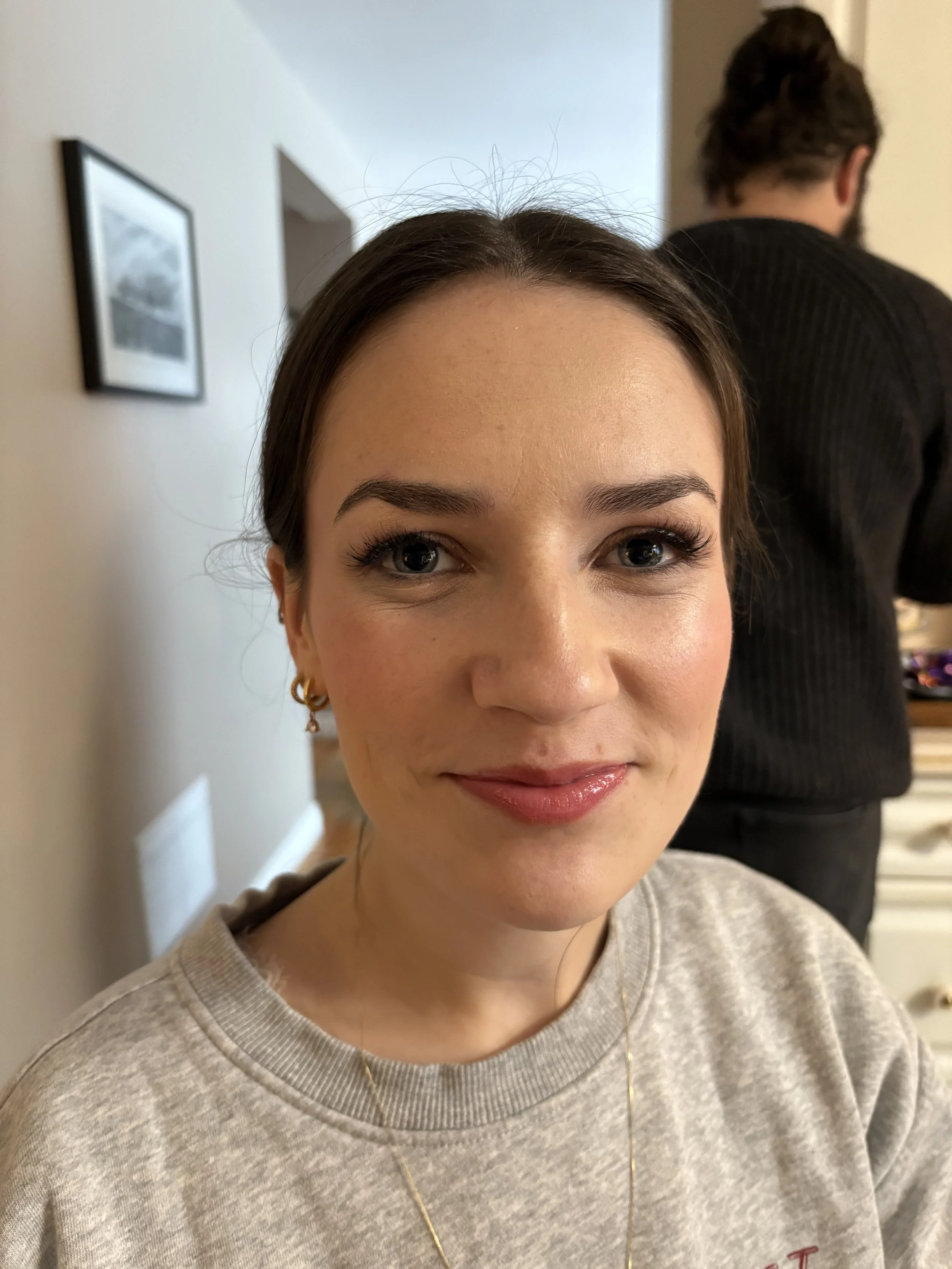 A woman with light skin and brown hair smiling at the camera, wearing makeup, earrings, and a light-colored sweatshirt in an indoor setting, with a blurred man in the background.