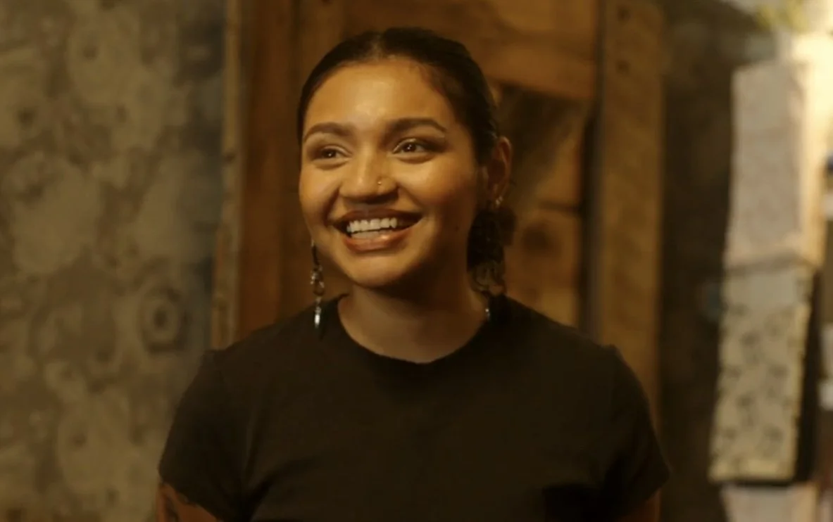 A woman with dark hair, earrings, and a nose piercing, smiling while looking to the side, in a warmly lit setting with stone and wood background.
