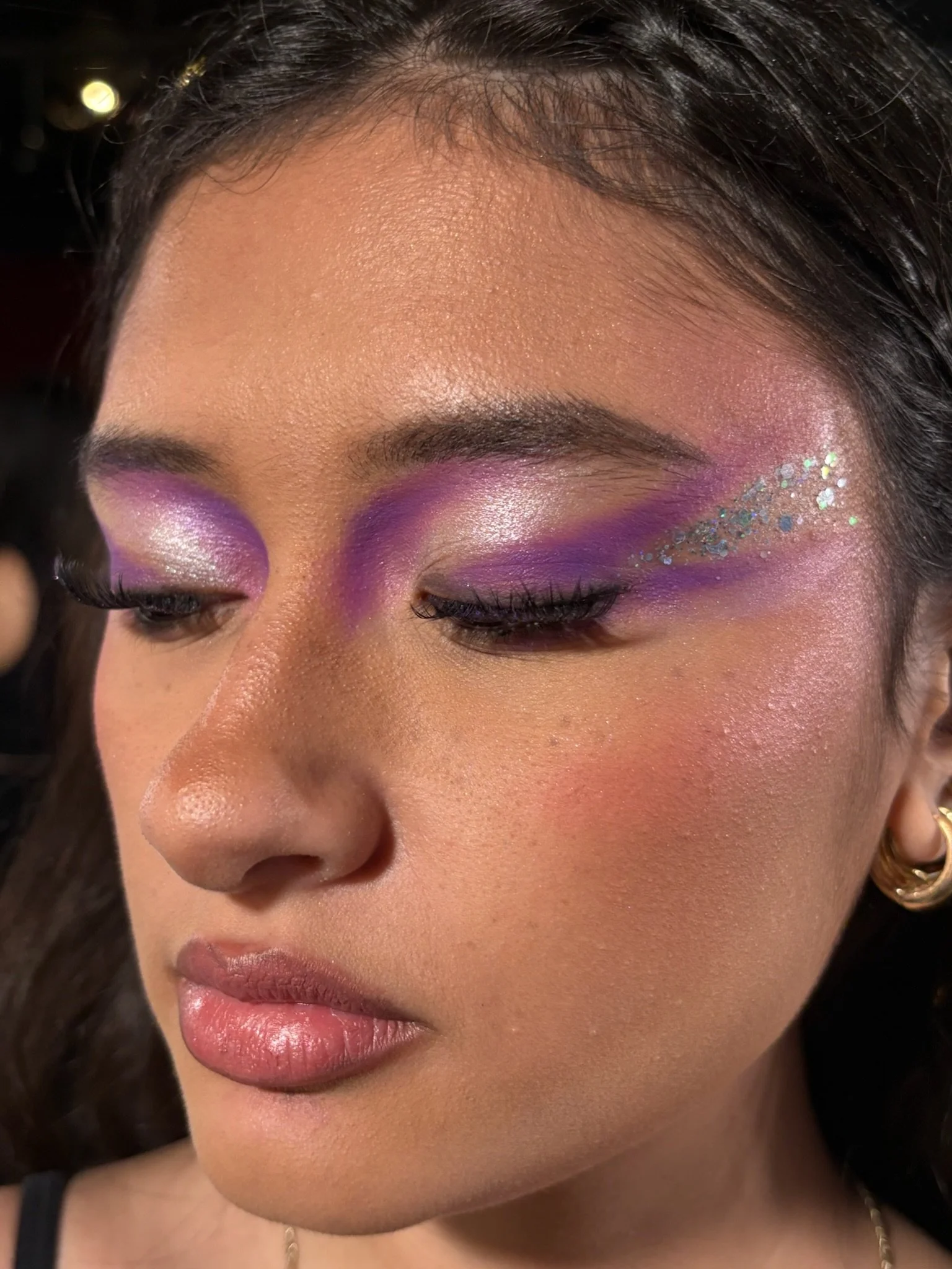 Close-up of a woman with colorful makeup, purple eyeshadow, silver shimmer, and glitter on her eyelids and cheekbones.