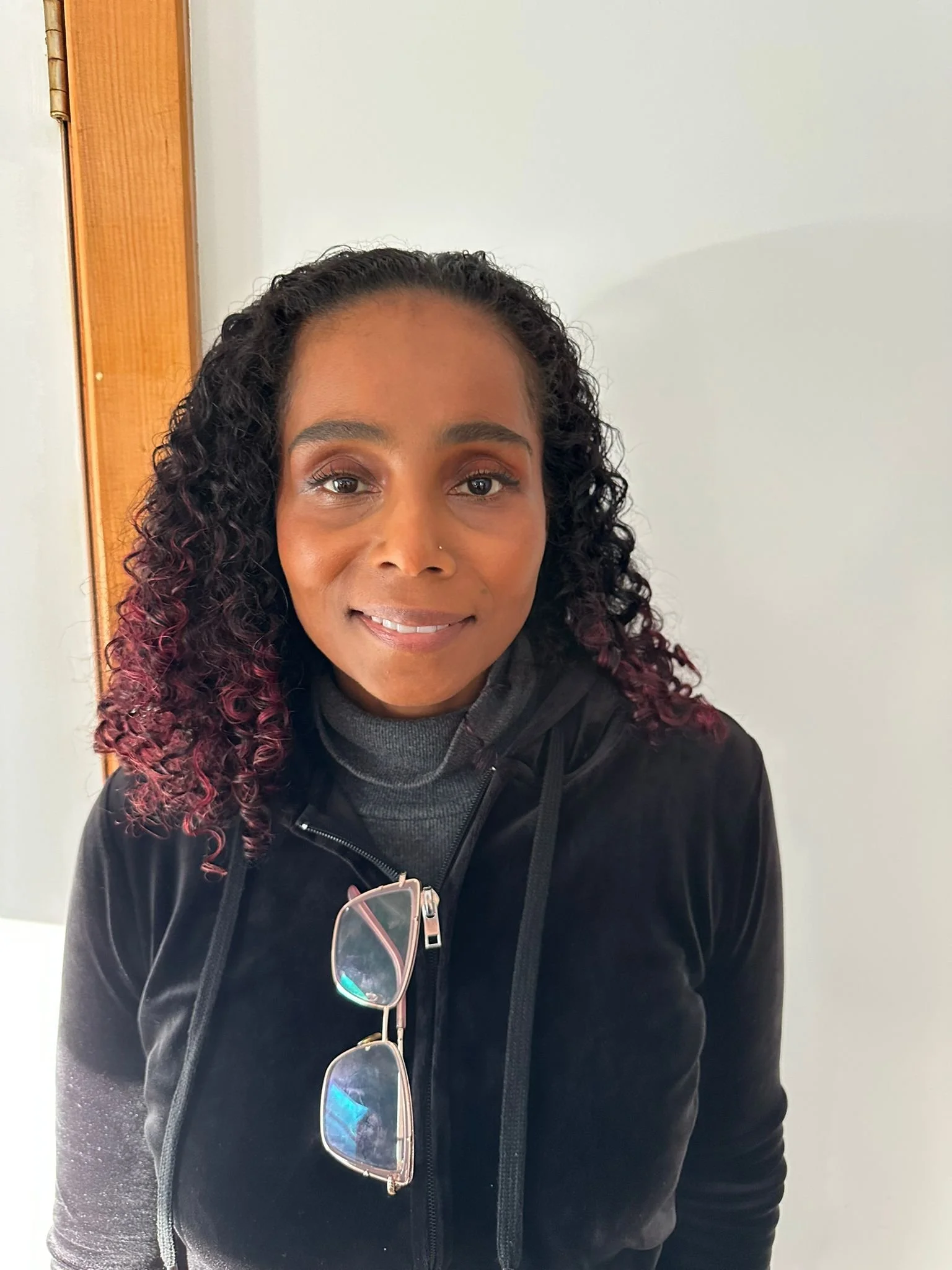 A woman with curly dark hair with burgundy tips, wearing a black hoodie and sunglasses hanging from the hoodie, smiling indoors against a plain wall.